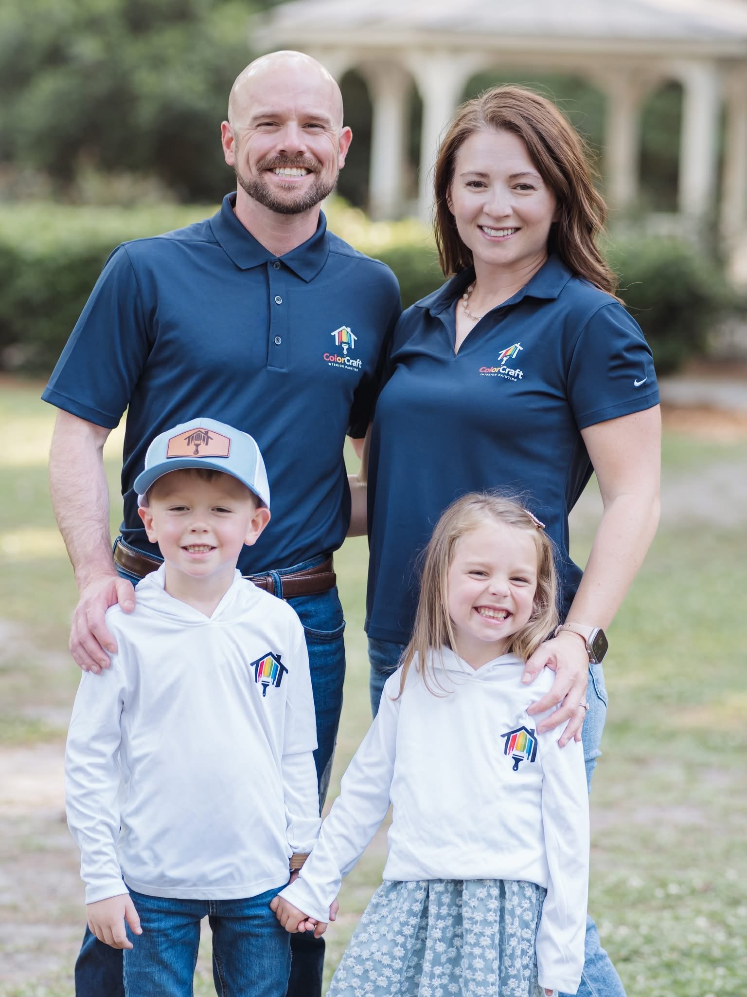 A family of five standing outdoors on a grassy area with a bridge in the background. They are all smiling and wearing matching navy blue shirts with a 'ColorCraft' logo. The children are holding hands, and everyone is looking at the camera.
