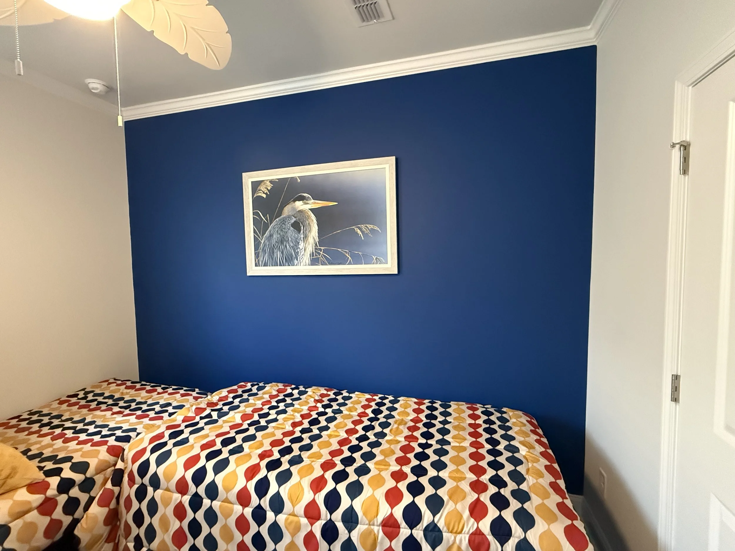 A bedroom with a bed covered in a colorful geometric patterned comforter. A blue accent wall features a framed picture of a heron bird standing among plants. The ceiling fan has a light with leaf-shaped blades.