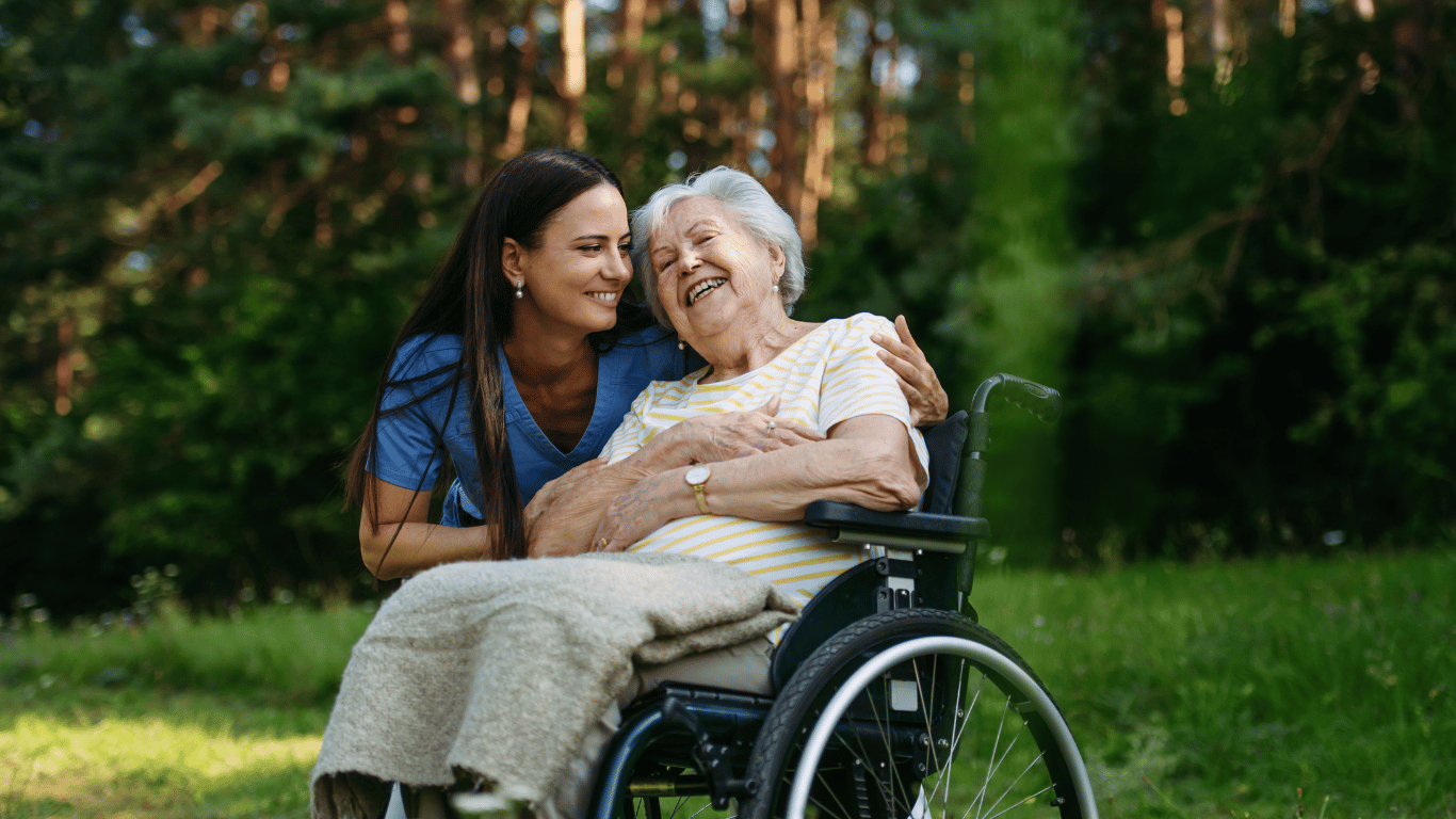 A young woman in medical scrubs hugging an elderly woman in a wheelchair outdoors surrounded by trees and greenery, both smiling and appearing joyful.