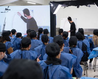Un profesor enseña a un grupo de estudiantes en una aula con pantalla grande y pizarras, todos usando batas azules.