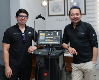 Dos hombres en un consultorio dental, uno con gafas y el otro con gafas de sol en la cabeza, posando junto a un equipo dental digital y una pantalla de ordenador mostrando una radiografía dental.