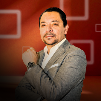 Hombre con barba y bigote poseando con los brazos cruzados, vestido con saco gris y camisa blanca, en un fondo de color rojo con cuadros blancos.