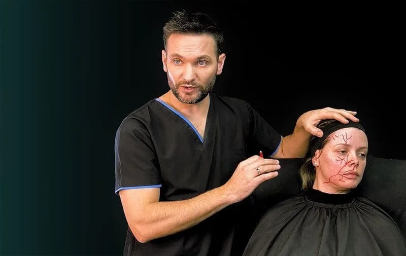 Un hombre vestido con una bata médica realiza un procedimiento de maquillaje de efectos especiales en el rostro de una mujer, creando venas y venas racemosas en su rostro.
