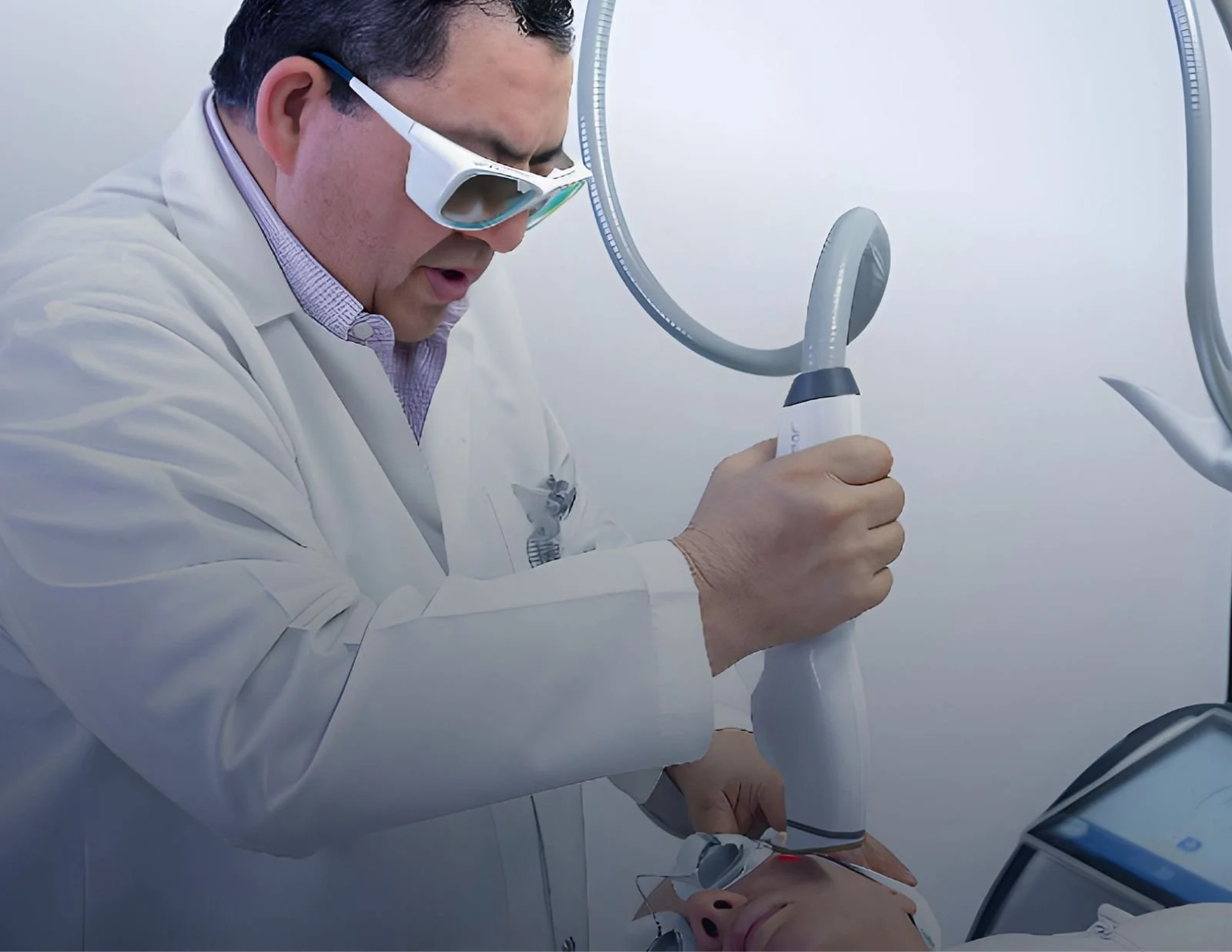Médico realizando un procedimiento en un paciente usando equipo de alta tecnología, con gafas protectoras y en un ambiente clínico.