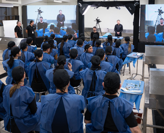 Estudiantes en uniforme azul asistiendo a una clase virtual con un instructor en una pantalla grande.