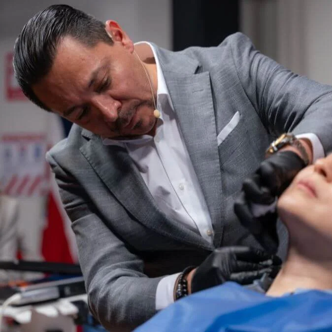 Un hombre con traje gris realiza un procedimiento médico en un rostro de una persona que está acostada, en un entorno clínico.