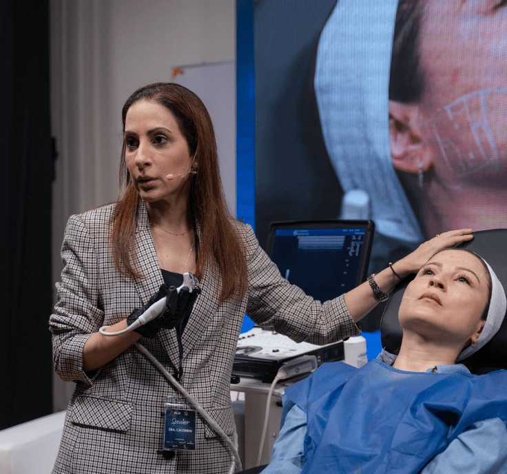 Una doctora realiza un procedimiento médico en una paciente en un entorno clínico, con equipo médico y una pantalla en el fondo.