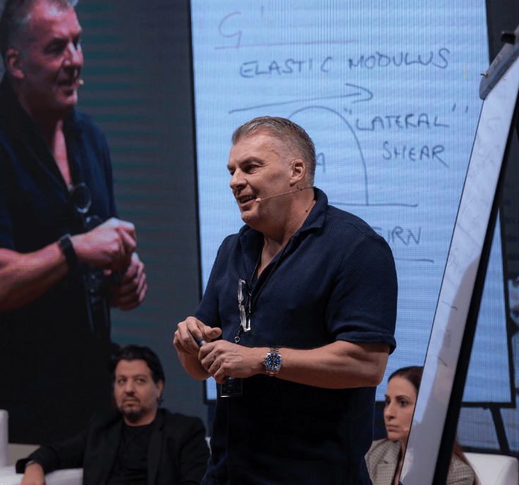 Hombre en una conferencia o taller, hablando frente a una pizarra con notas y diagramas, con varias personas sentadas en el fondo.