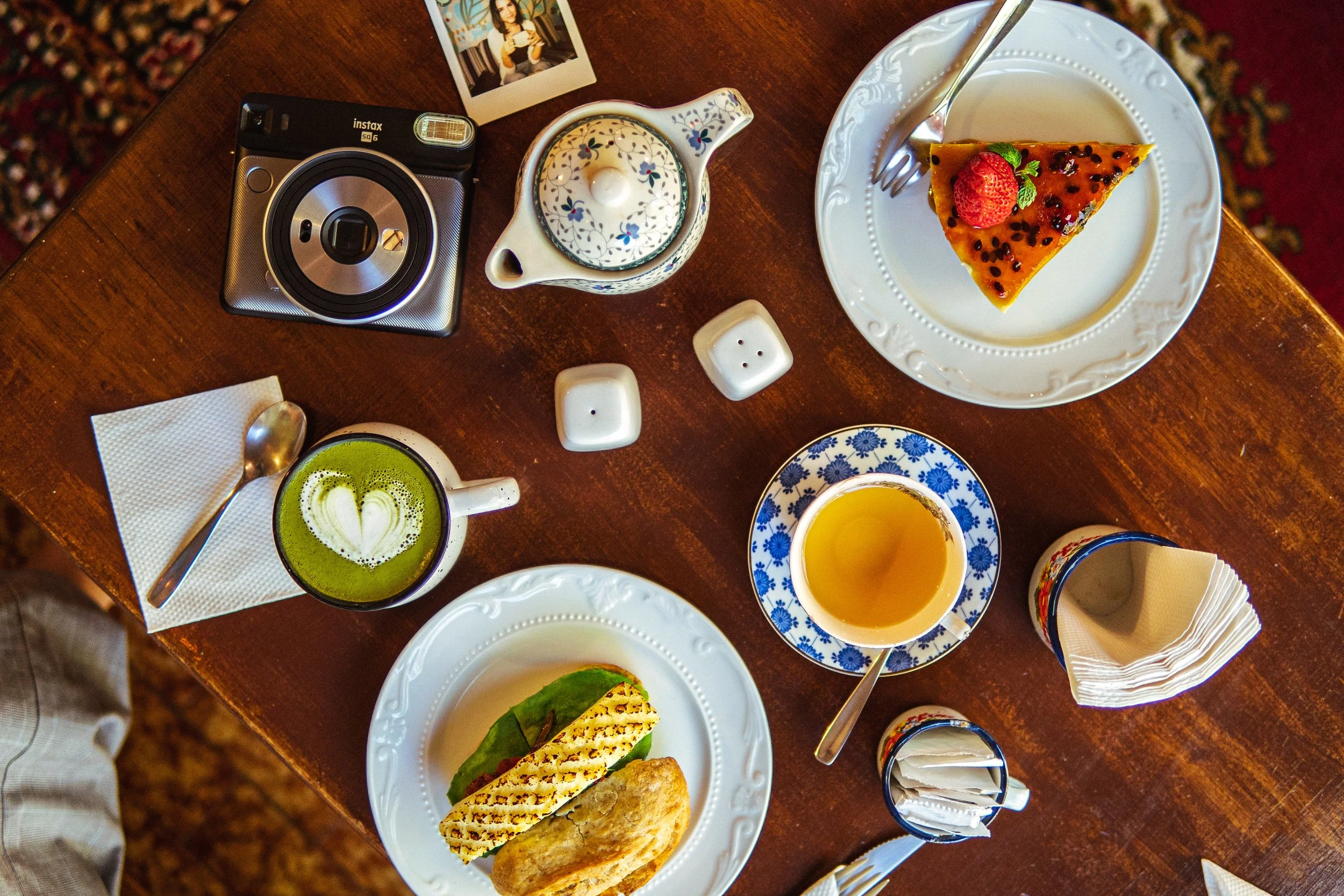 A wooden table set with various breakfast items including coffee, tea, a slice of berry-topped cake, a sandwich with grill marks, cutlery, a Polaroid photo, a camera, salt and pepper shakers, and some napkins.