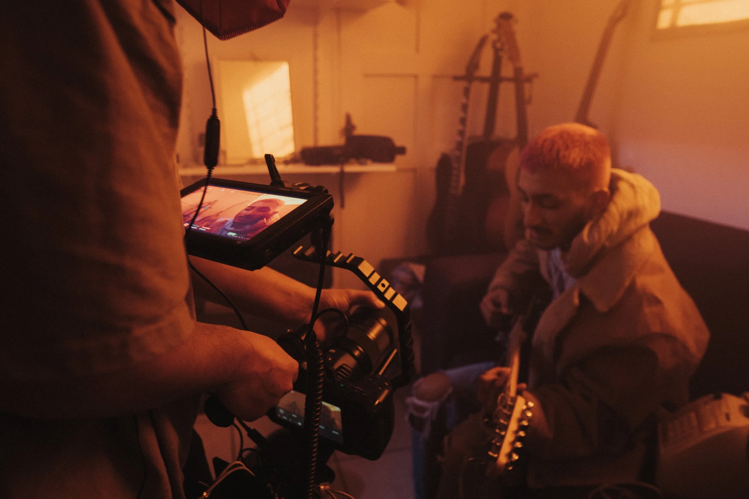 Behind-the-scenes photo of a film or photo shoot with a person in a beige hoodie, sunglasses, and colorful hair, sitting in a dimly lit room, while crew members operate camera equipment.