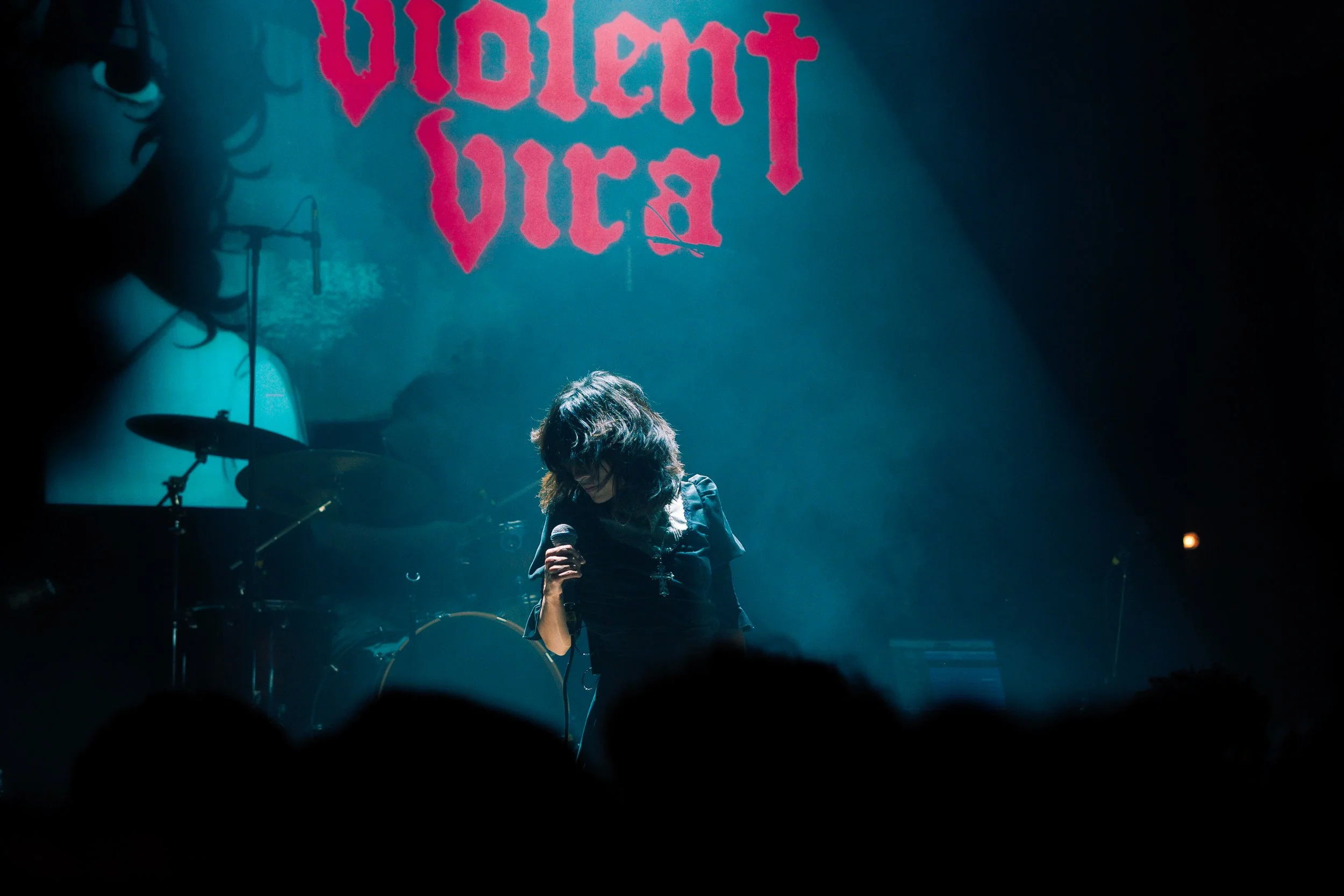 A woman with curly hair holds a microphone on stage during a concert, with a drum set and dark background behind her. A large, red, gothic-style text sign reads 'Violet Vault' on a blue-green wall.