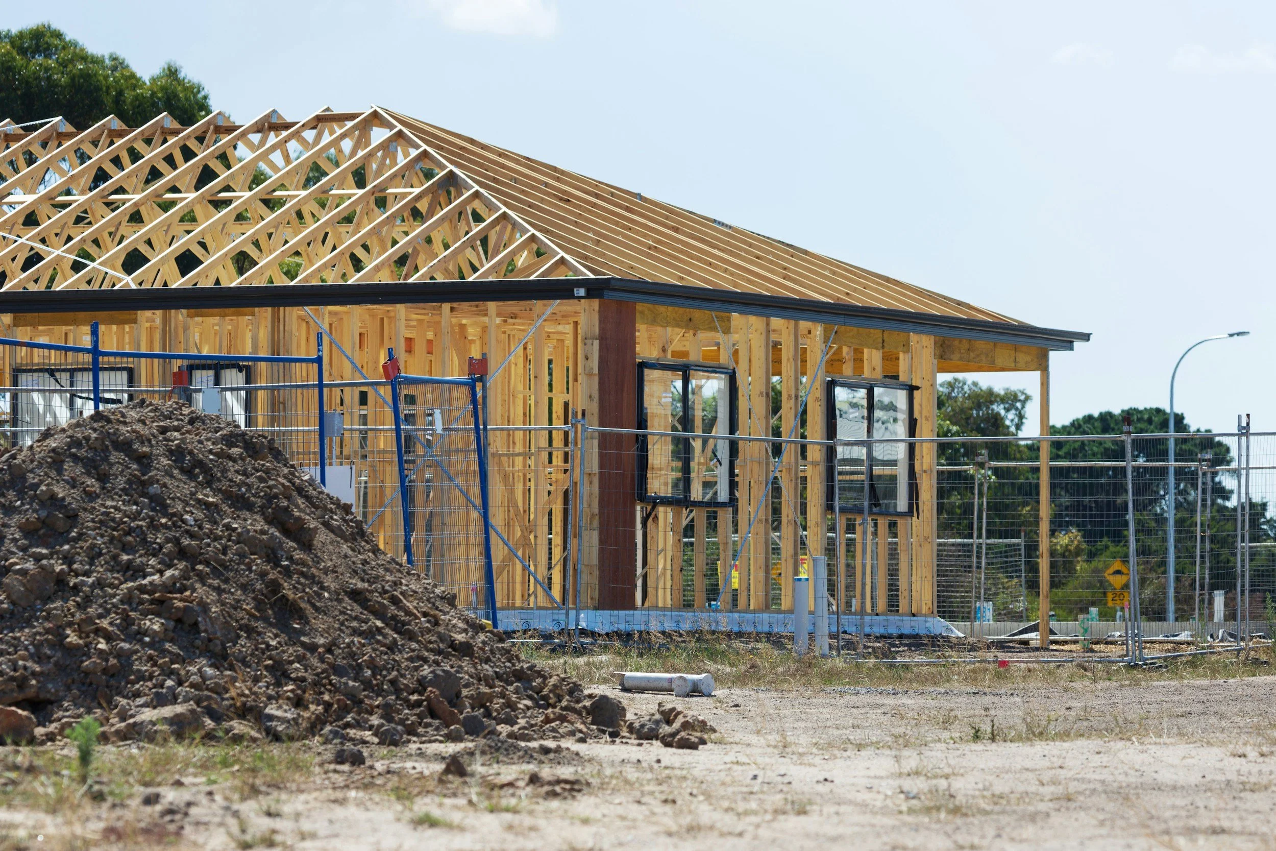 Construction site with a partially built wooden house, surrounded by fencing, with a pile of dirt in the foreground and trees in the background.