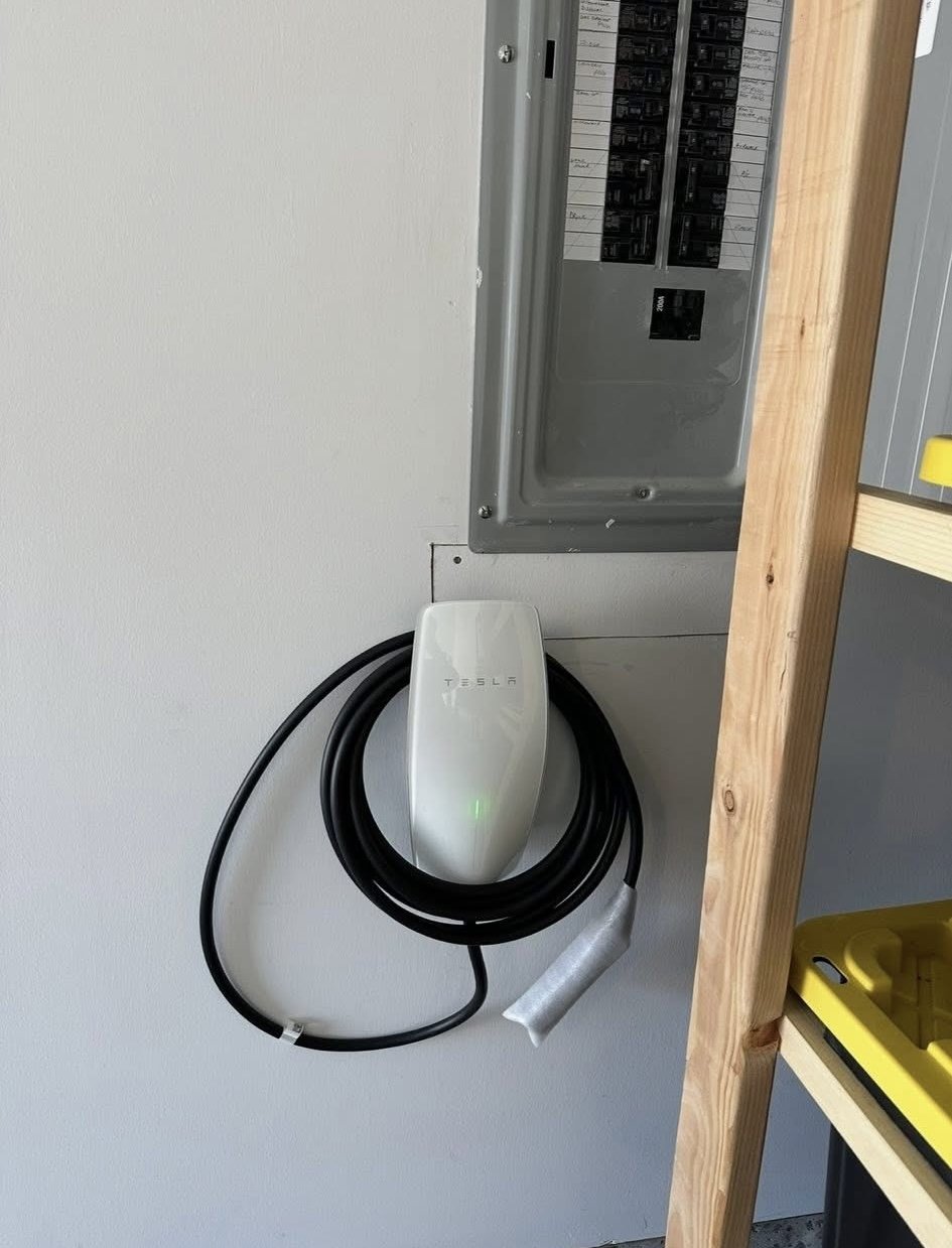 Tesla electric vehicle charger attached to a white wall, with a black charging cable coiled around it.