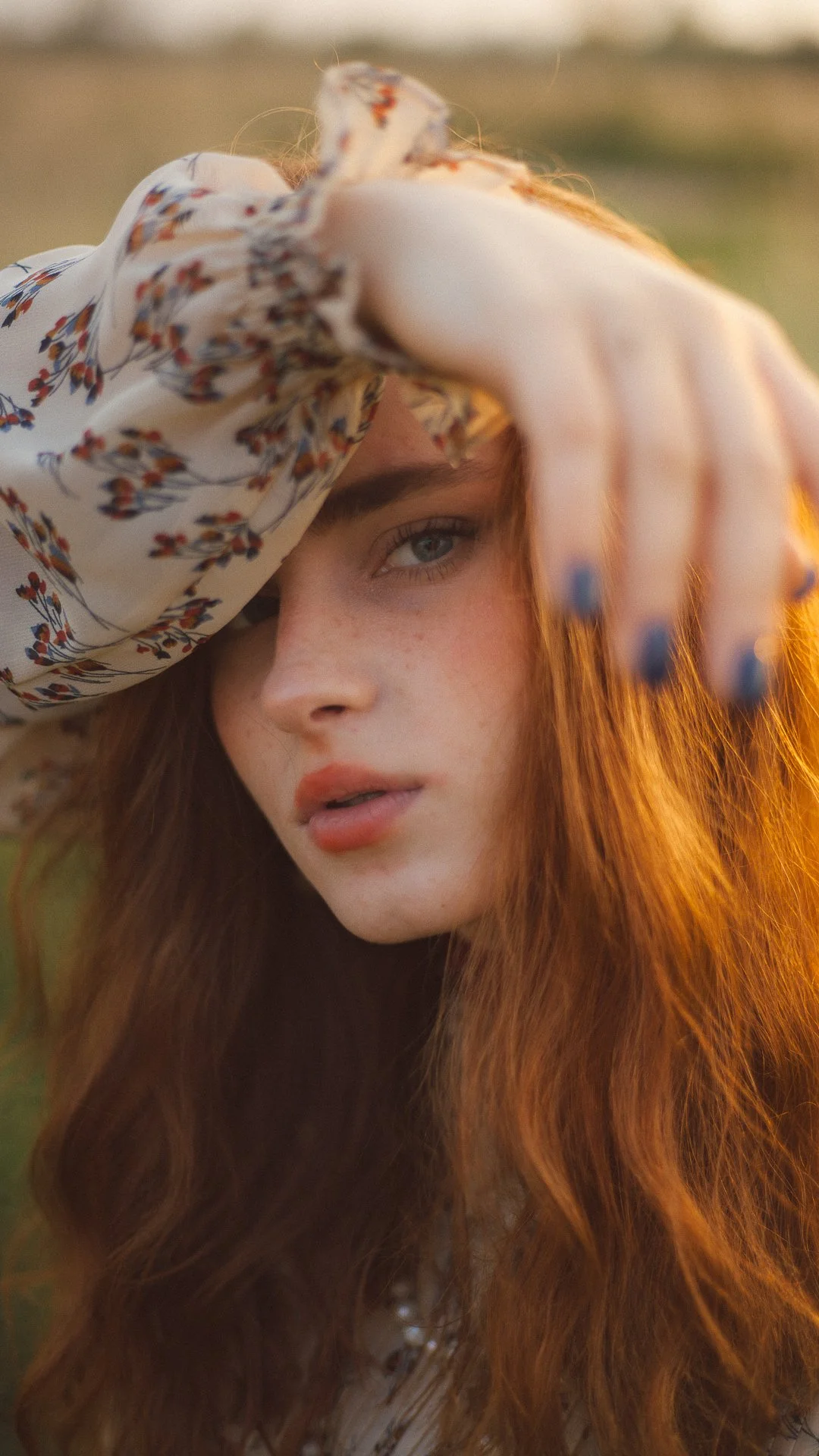 A close-up of a young woman with long red hair and blue eyes, holding her hand up to her forehead outdoors during golden hour, wearing a floral blouse with a transparent fabric.