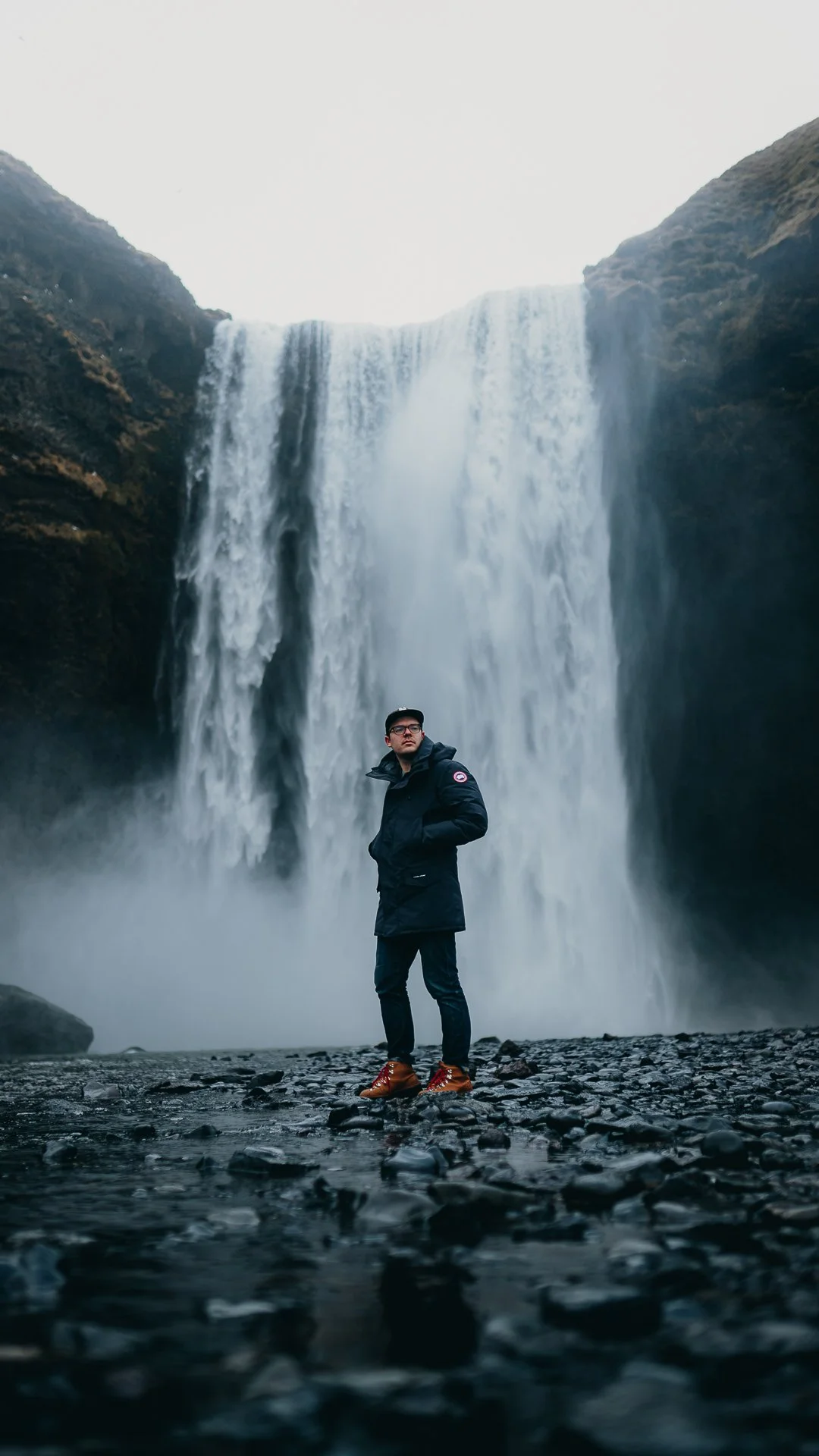 A person standing on rocky ground in front of a large waterfall surrounded by mountains.