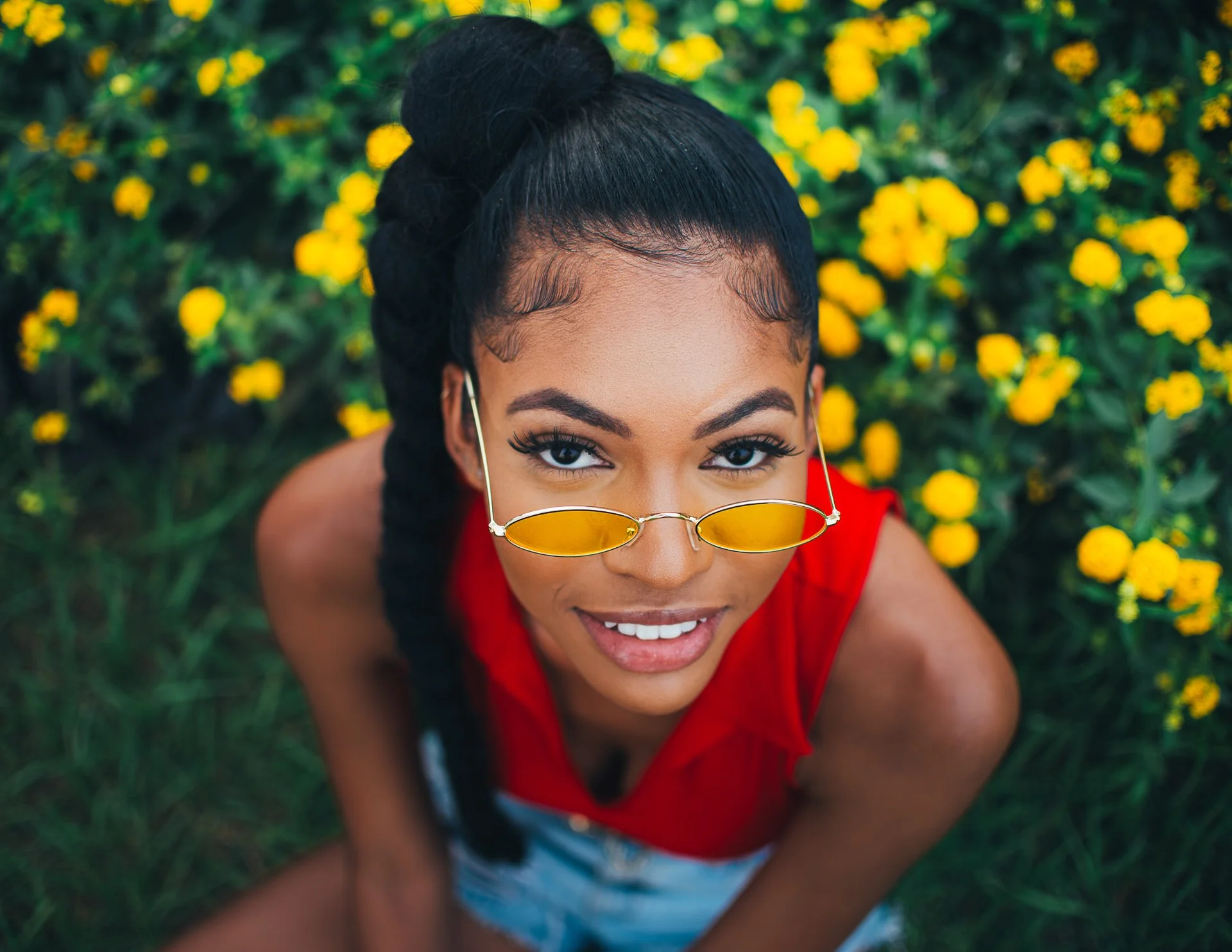 A young woman with dark hair in a bun and sunglasses, smiling and looking up at the camera, surrounded by yellow flowers.