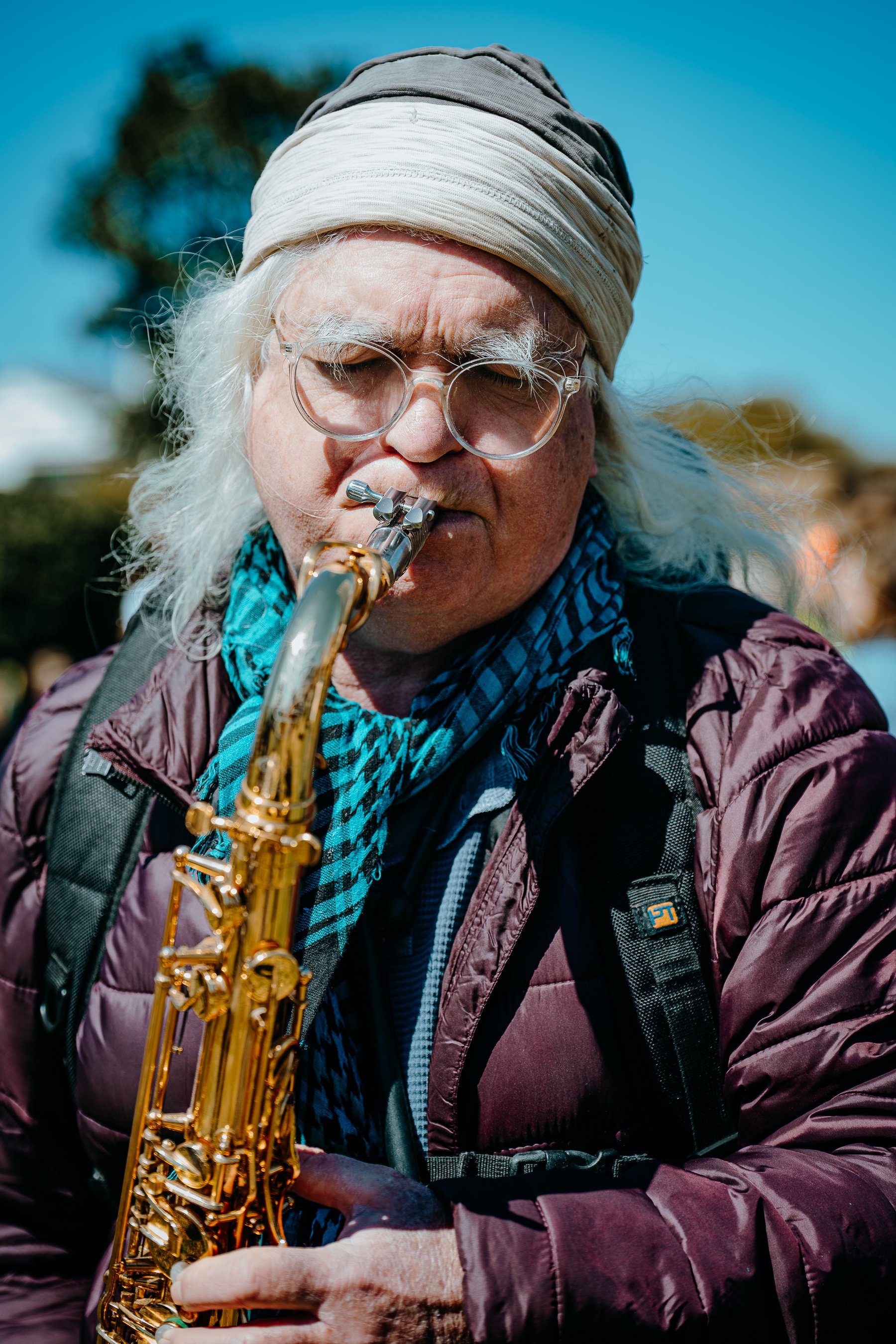 An elderly man with white hair, wearing glasses, a beige headscarf, and a purple jacket, playing a gold-colored saxophone outdoors on a sunny day.