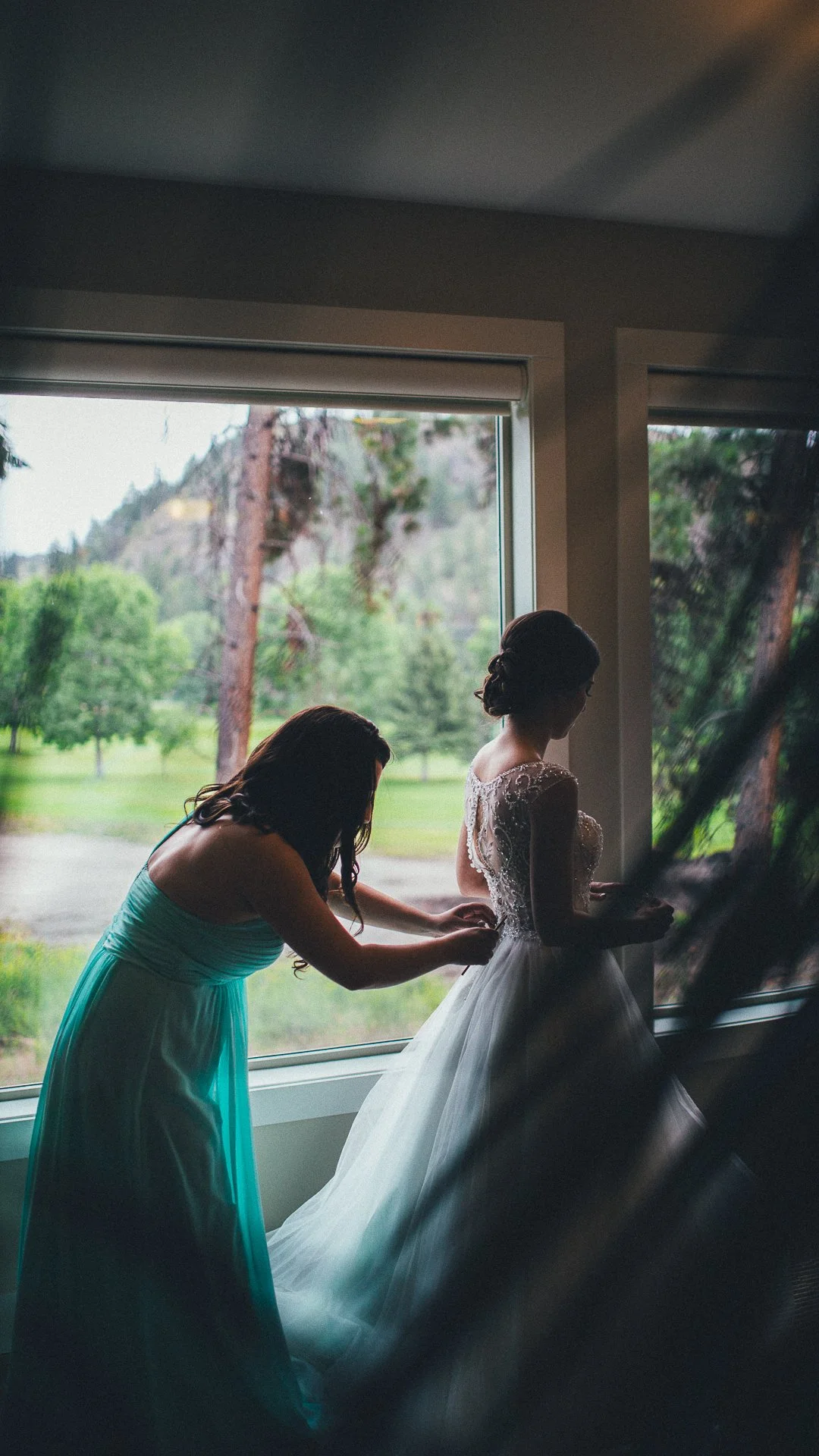 A woman in a wedding dress preparing for her wedding while another woman assists her, standing by a large window with scenic outdoor trees and mountains.