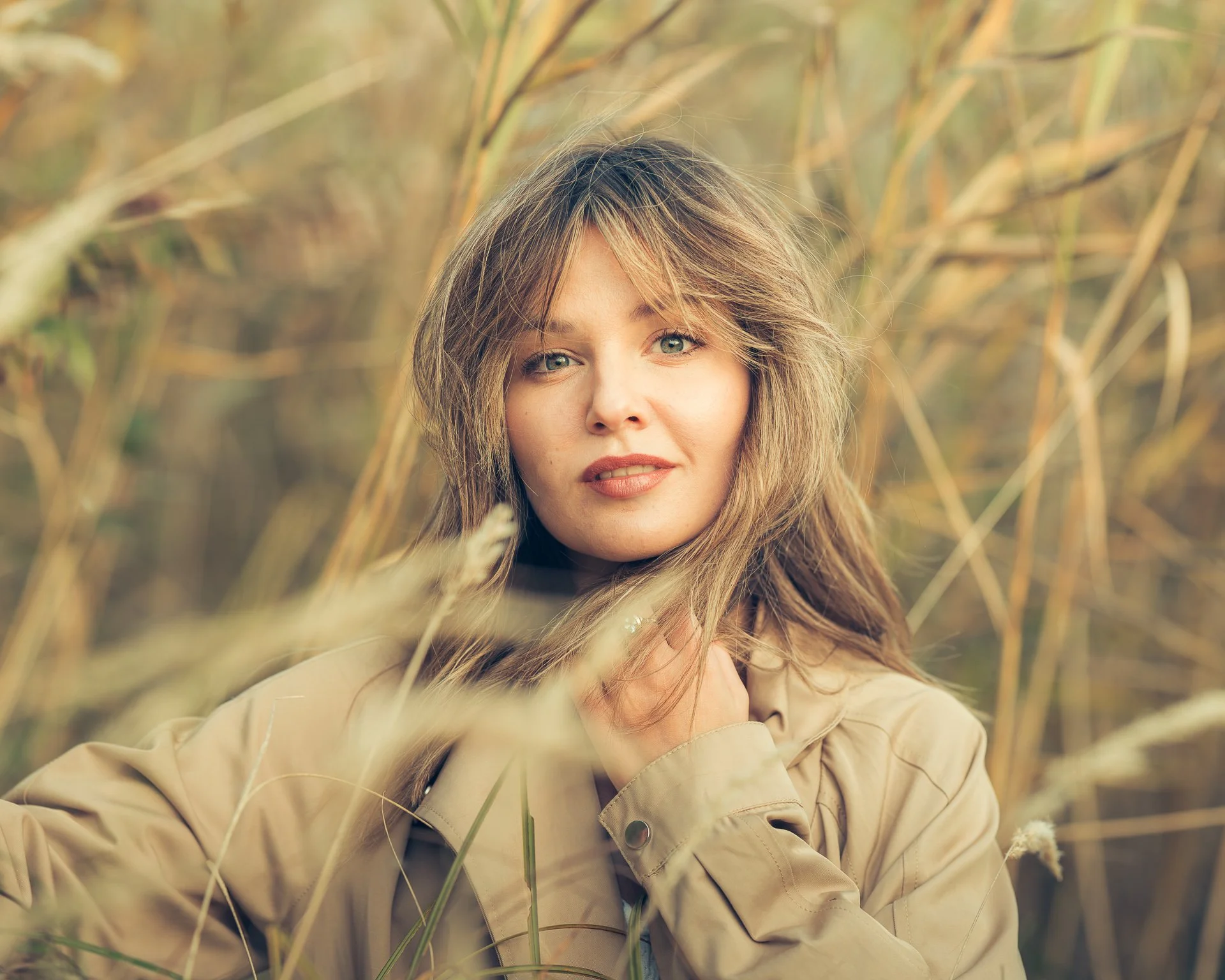 A woman with wavy light brown hair and blue eyes outdoors among tall grasses