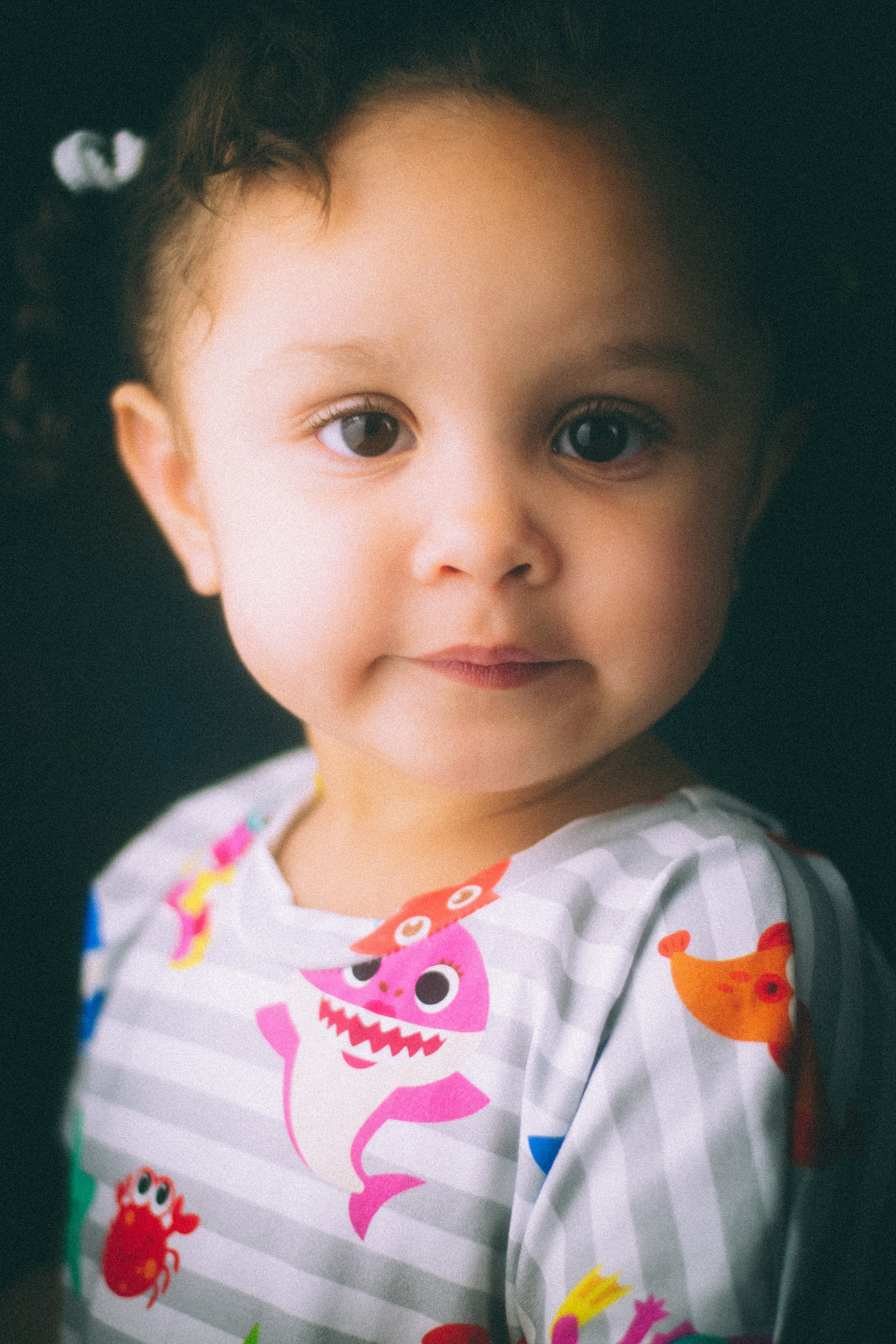 Close-up of a young girl with big eyes, wearing a colorful shirt with cartoon monsters and animals, smiling softly against a dark background.