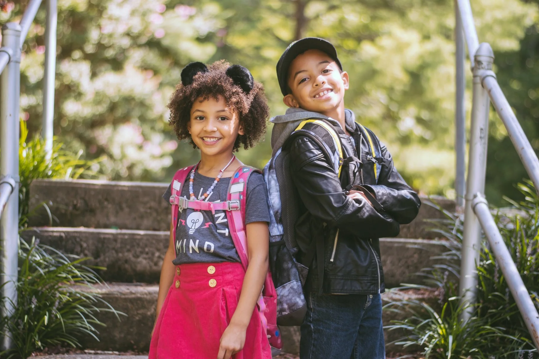 Two young children, a girl and a boy, standing outdoors on a staircase, smiling. The girl has curly hair with hair accessories and a pink backpack, wearing a gray T-shirt and a pink skirt. The boy has short hair, wearing a black leather jacket, a gra