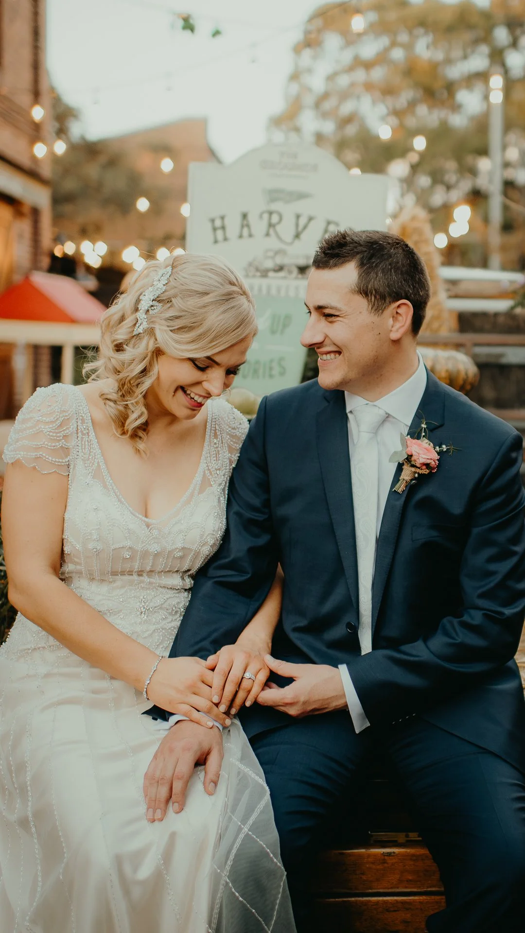 A bride and groom sitting close together outdoors, smiling and laughing. The bride is wearing a white wedding dress with decorative beading and a headband, and the groom is in a dark blue suit with a white shirt and tie. The bride's hand with a weddi