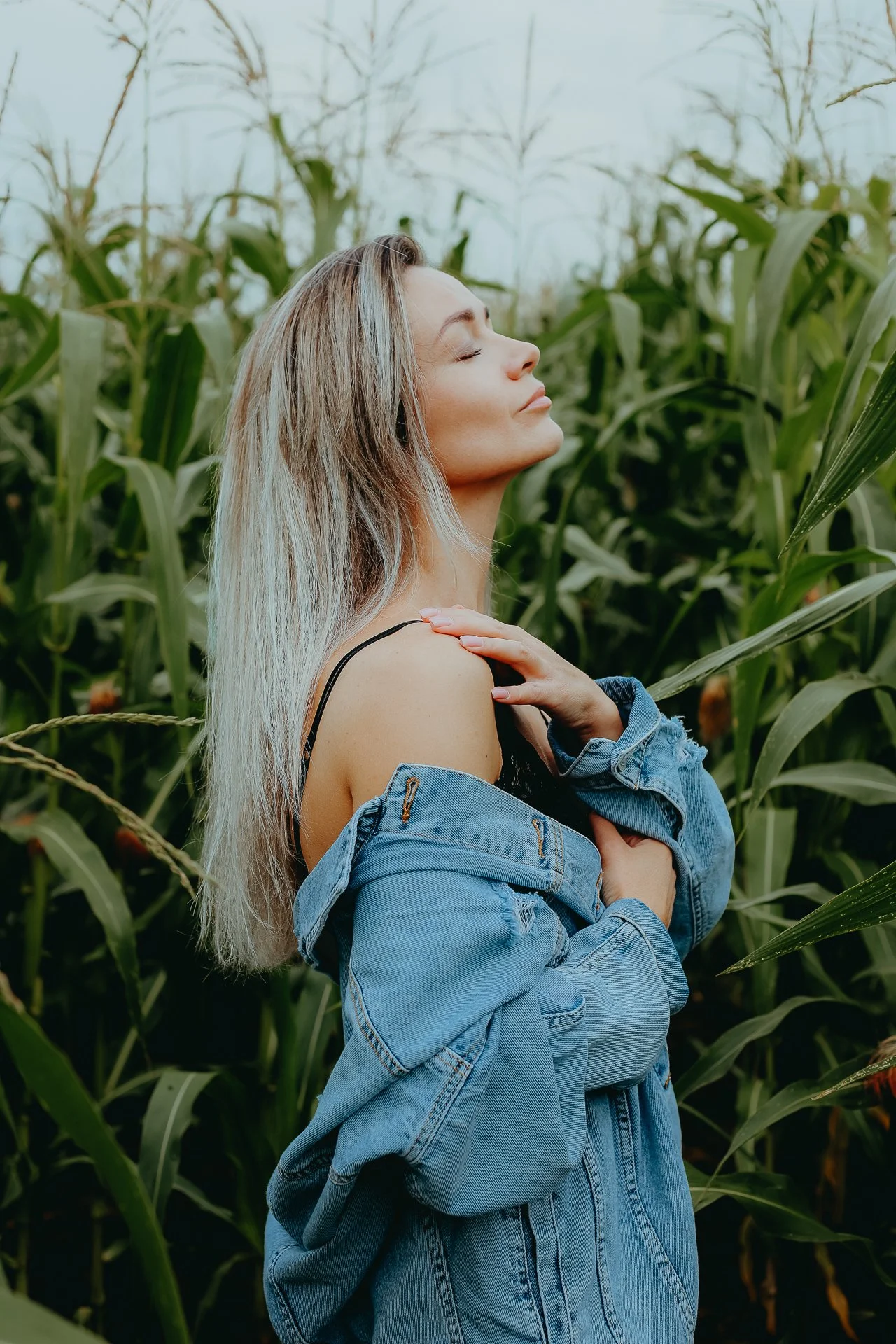 A woman with long blonde hair standing in a field of tall green corn plants, wearing a black top and a blue denim jacket with one shoulder exposed, with her eyes closed and face turned slightly upward.