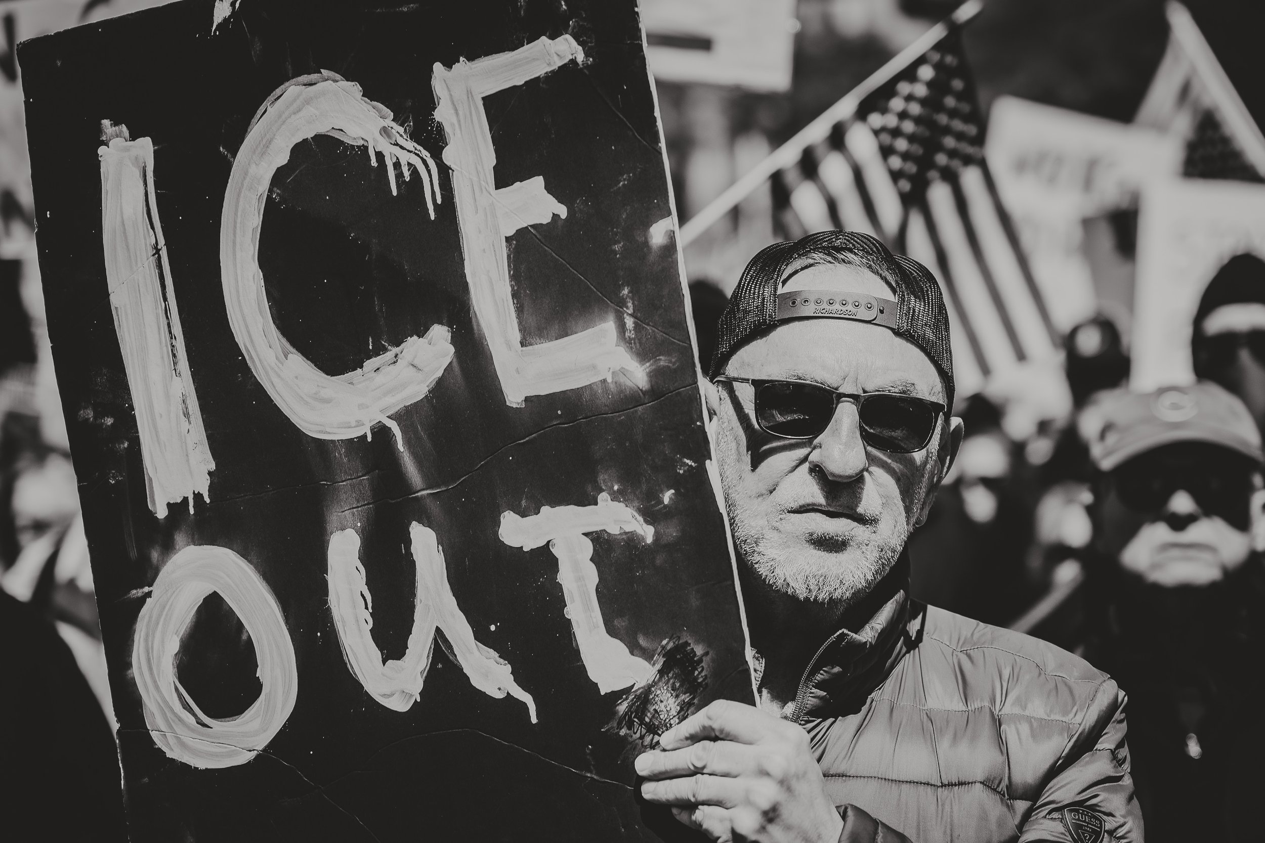 A man wearing sunglasses and a cap holding a sign that reads "ICE OUT" at a protest or demonstration with American flags and other people in the background