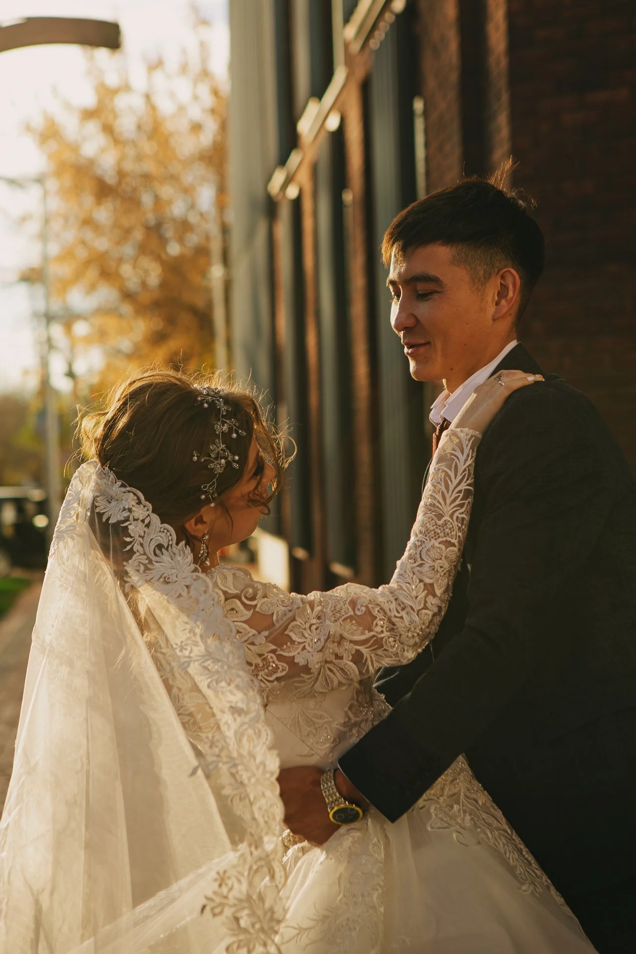 A bride and groom share a tender moment outdoors during sunset, with the bride in a lace wedding dress and the groom in a dark suit, standing close together.