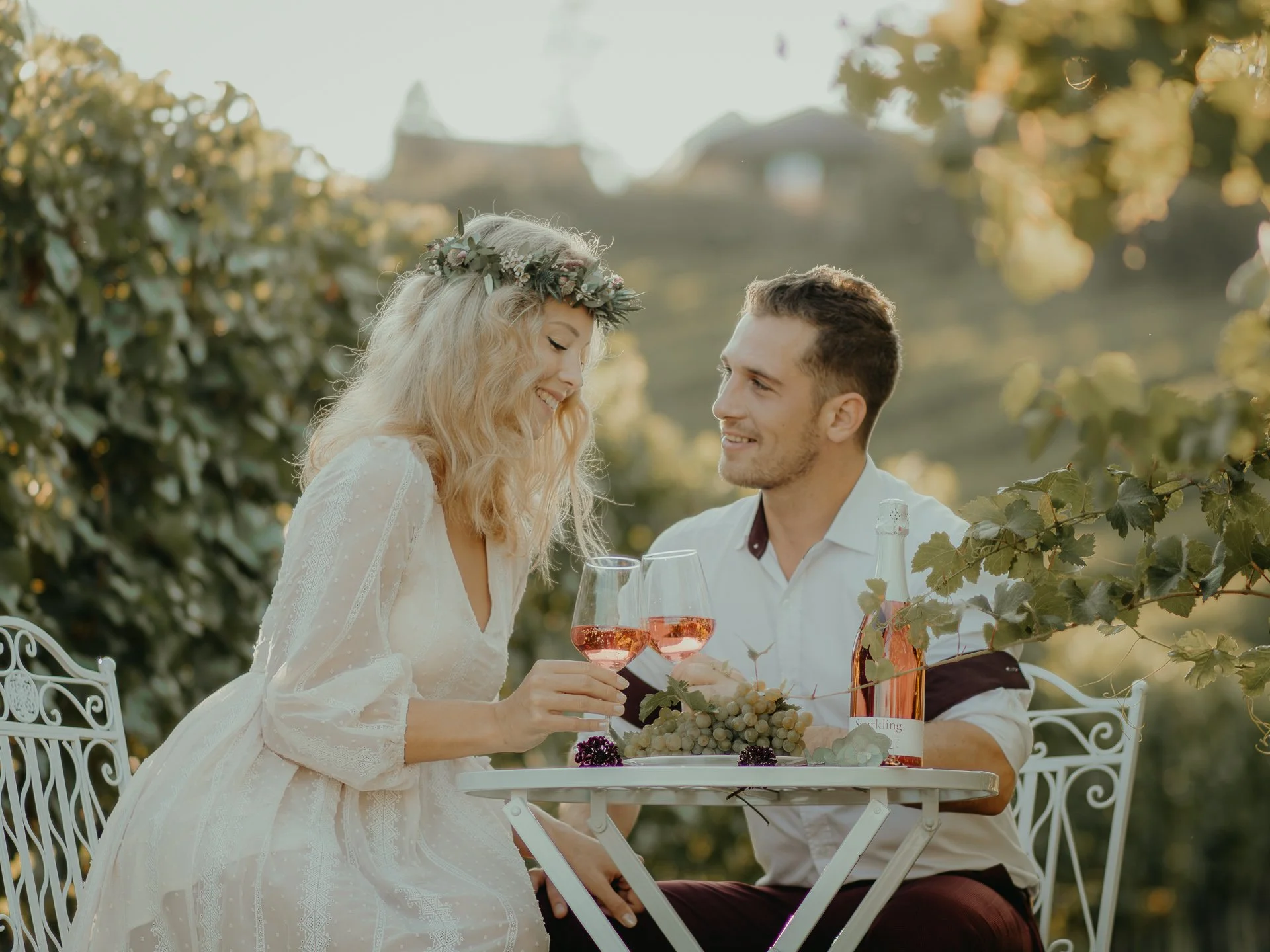 A woman with blonde hair wearing a lace dress and floral crown, and a man with short dark hair in a white shirt, are smiling and toasting with glasses of rosé wine at an outdoor setting with greenery and a blurred hillside in the background.