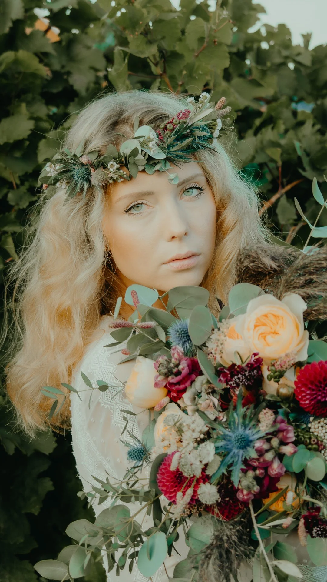 A woman with blonde, wavy hair and a floral crown, holding a large bouquet of colorful flowers, standing in front of green leafy bushes.