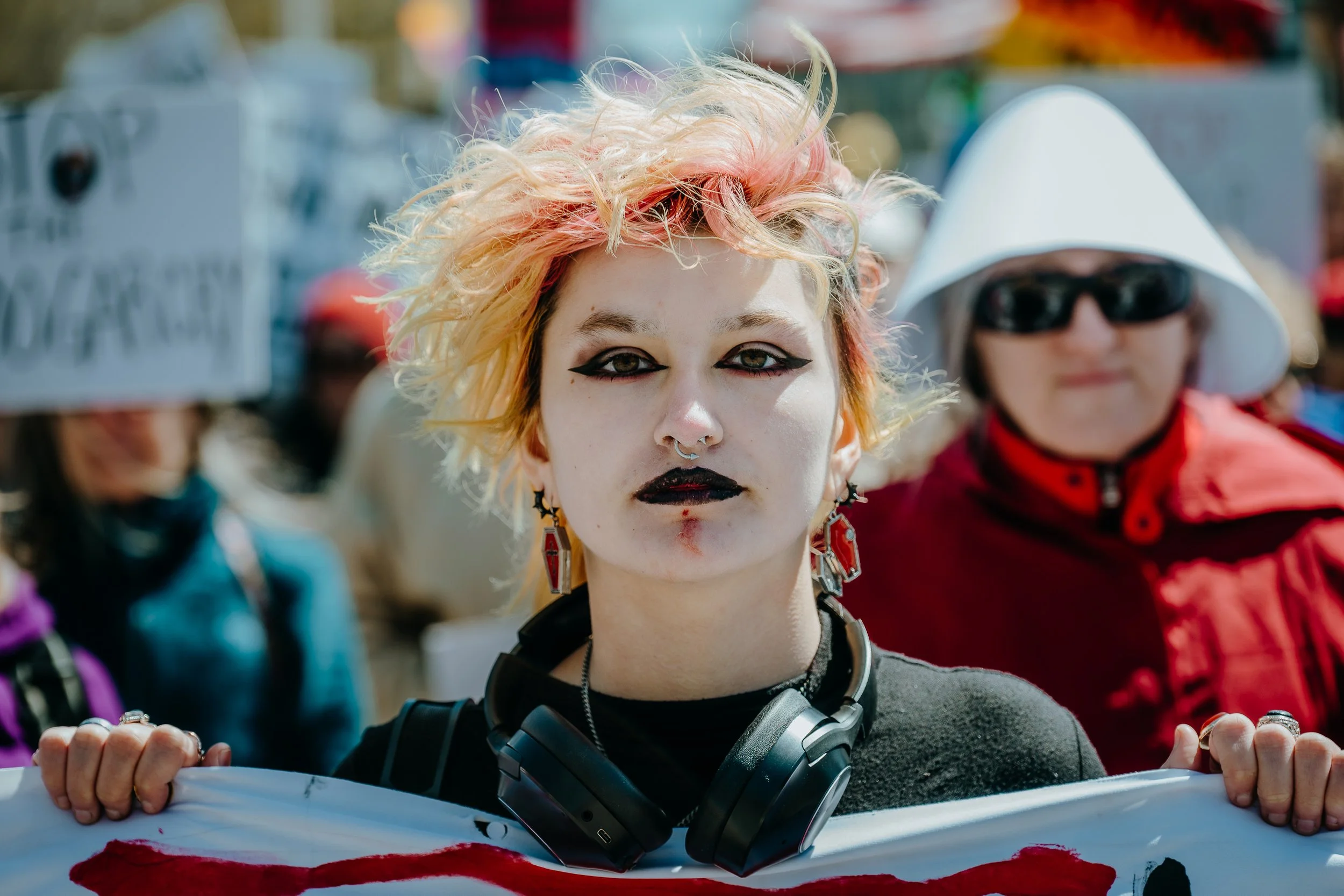A young woman with bleached, pink-tinted hair and gothic makeup holding a protest sign at a rally or demonstration, with other protesters in the background.