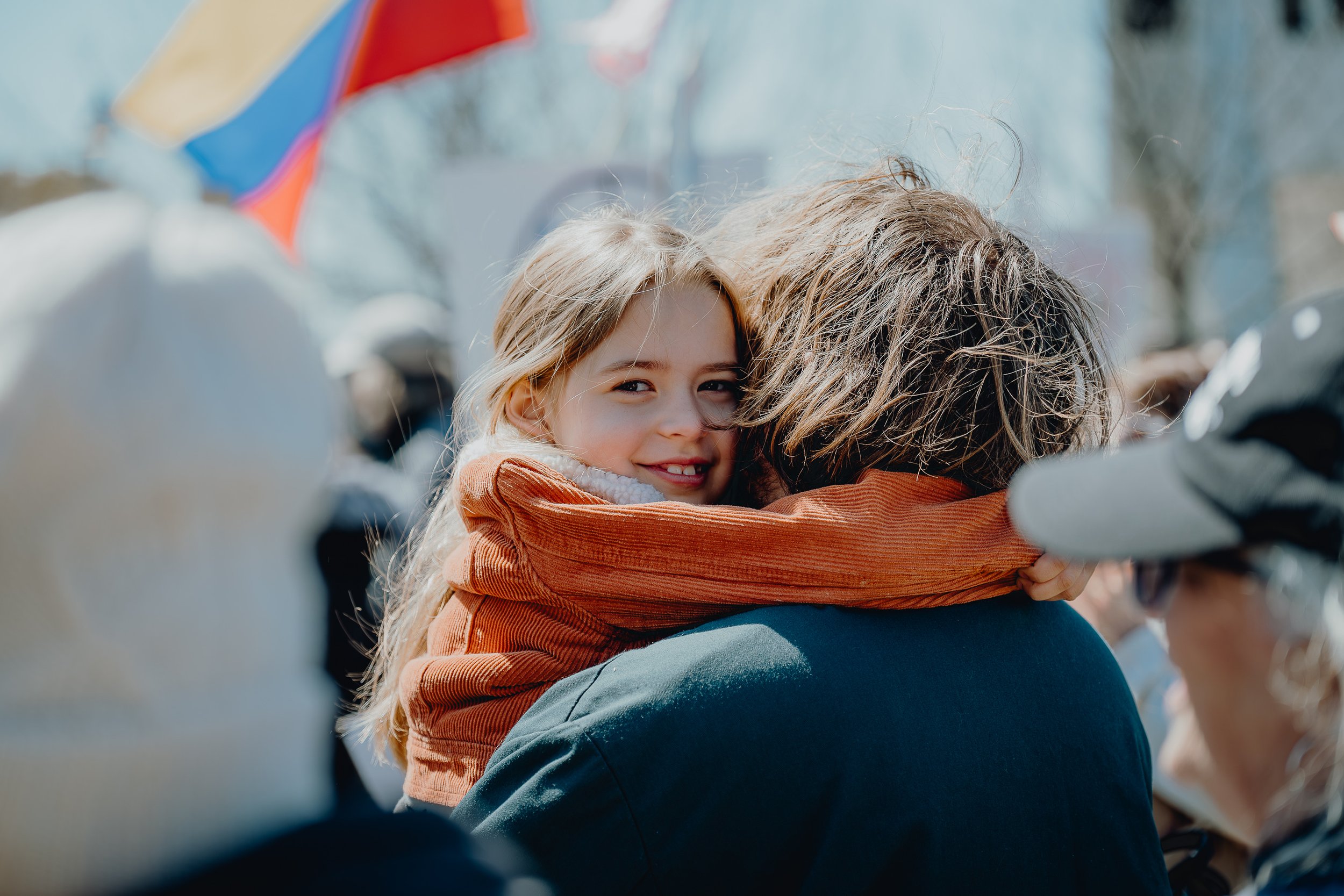 A young girl smiling and hugging an adult in a crowd outdoors on a sunny day, with a colorful flag in the background.