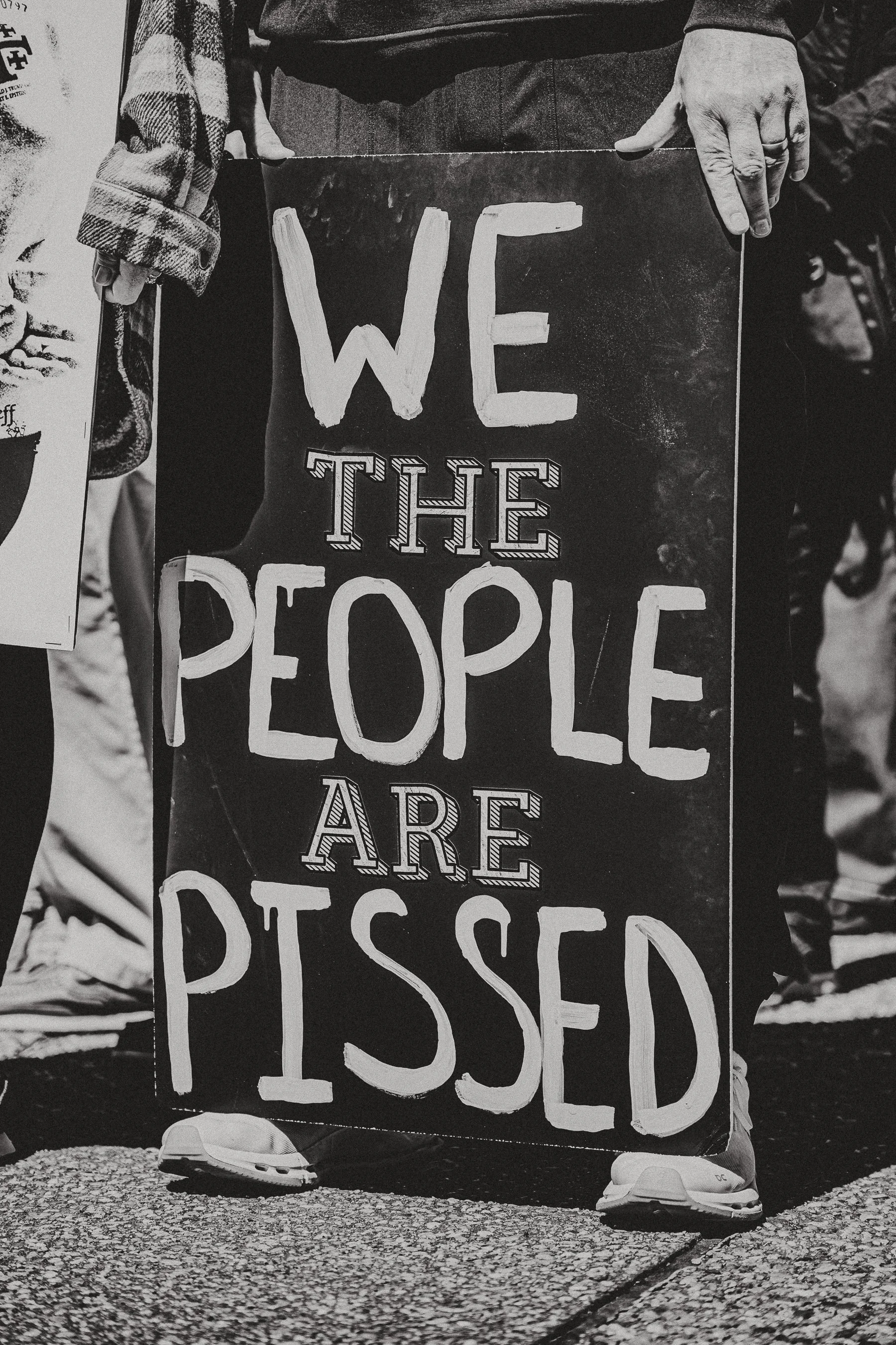 A person holding a sign that reads 'We the People Are Pissed' at a protest or rally. The image is in black and white.
