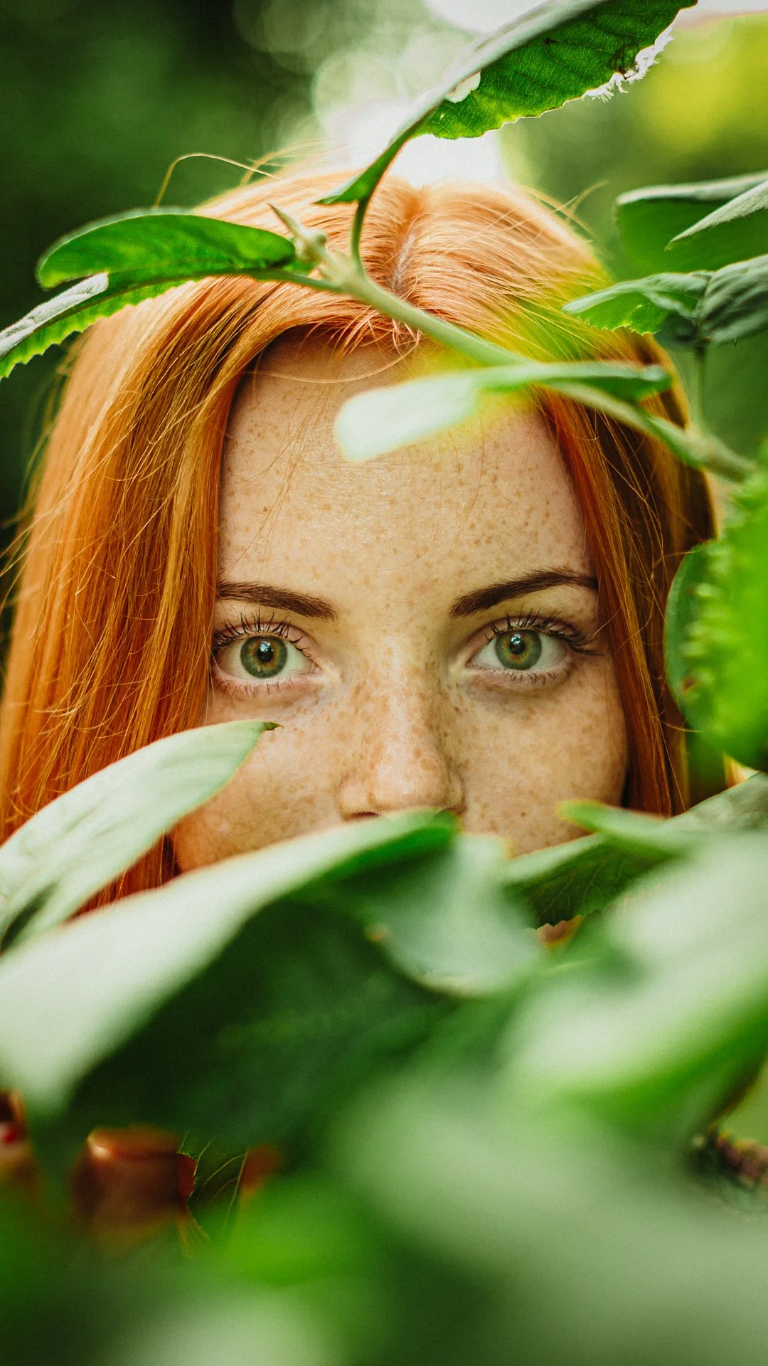 A close-up of a woman with green eyes and freckles, partially hidden behind green leaves, with her red hair visible.
