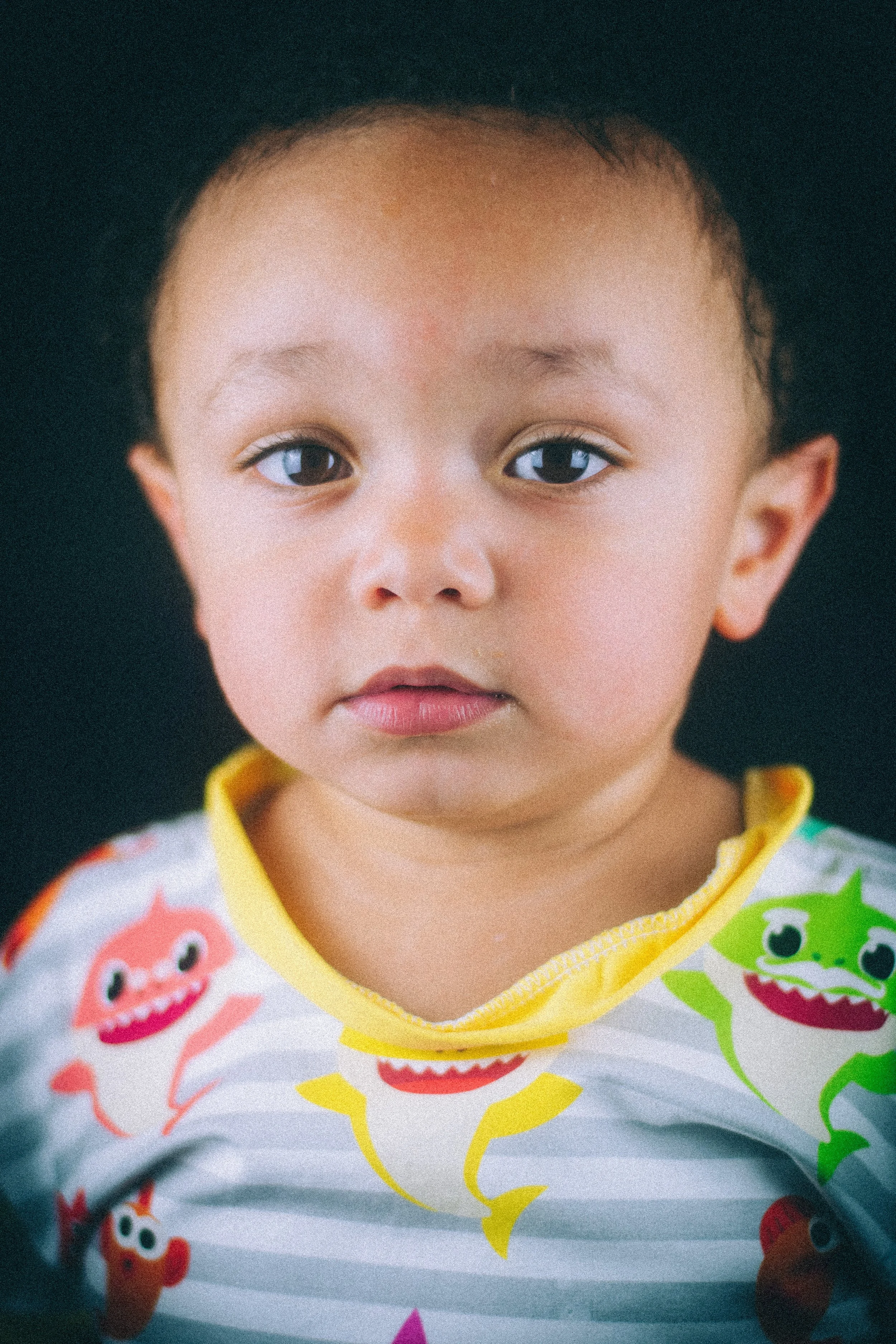 Close-up of a young child with light brown skin, short dark hair, and wearing a colorful shirt with cartoon monster prints, against a black background.
