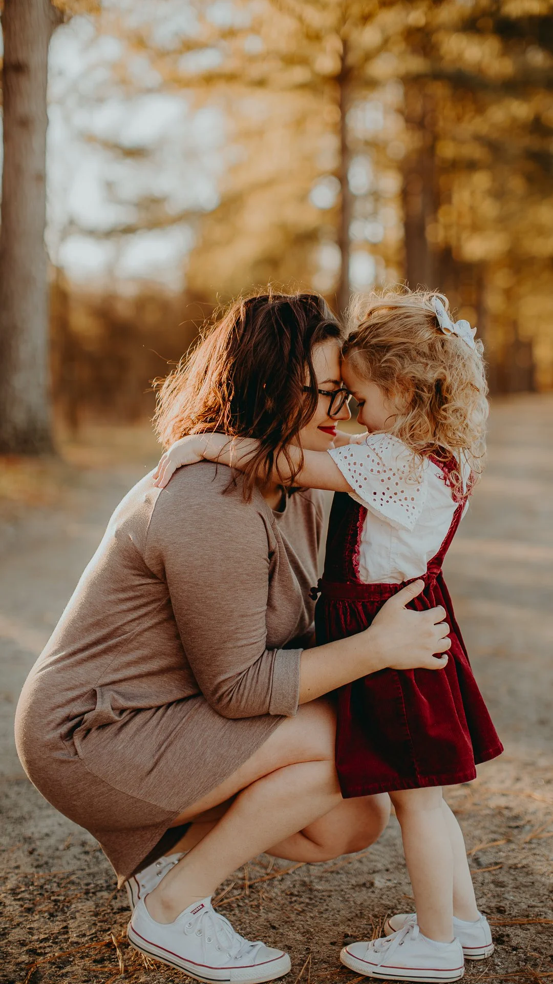 A woman crouches down to hug a young girl in an outdoor park during autumn, with trees in the background. Both are wearing white sneakers and have their foreheads touching, sharing an intimate moment.