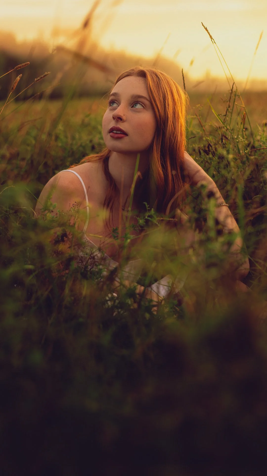 A young woman with red hair lies in a grassy field during sunset, gazing up with a thoughtful expression.