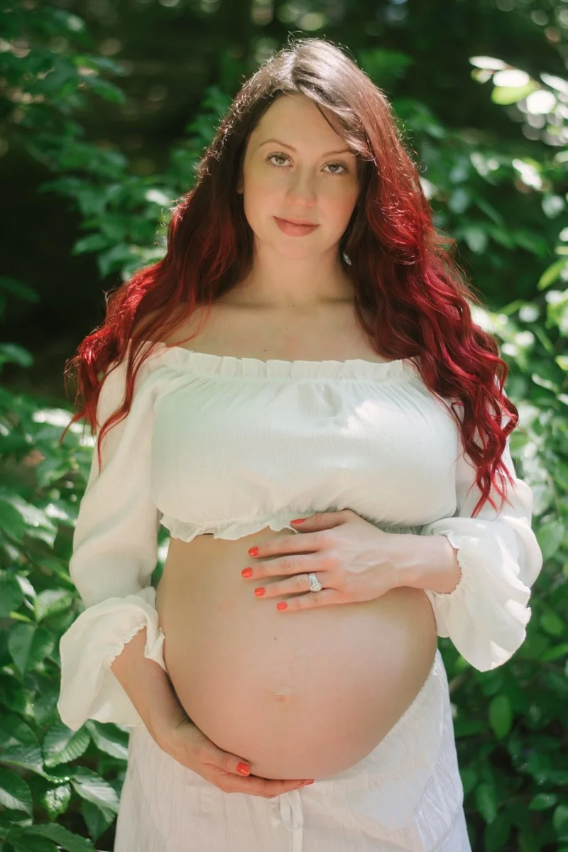 A pregnant woman with long red hair standing outdoors among green foliage, wearing a white off-the-shoulder top and skirt, holding her belly with one hand and touching her chest with the other.
