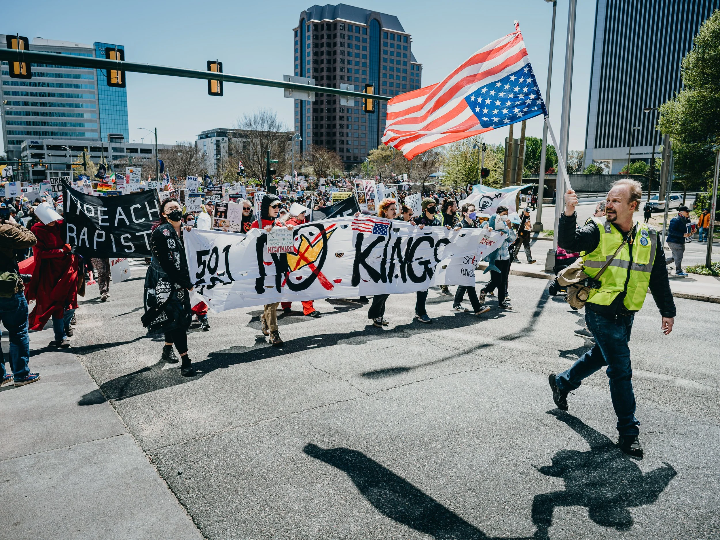 People protesting on a city street, holding signs and banners with messages against hate, racism, and violence. A man at the front waves an American flag. Modern buildings and traffic lights are visible in the background.