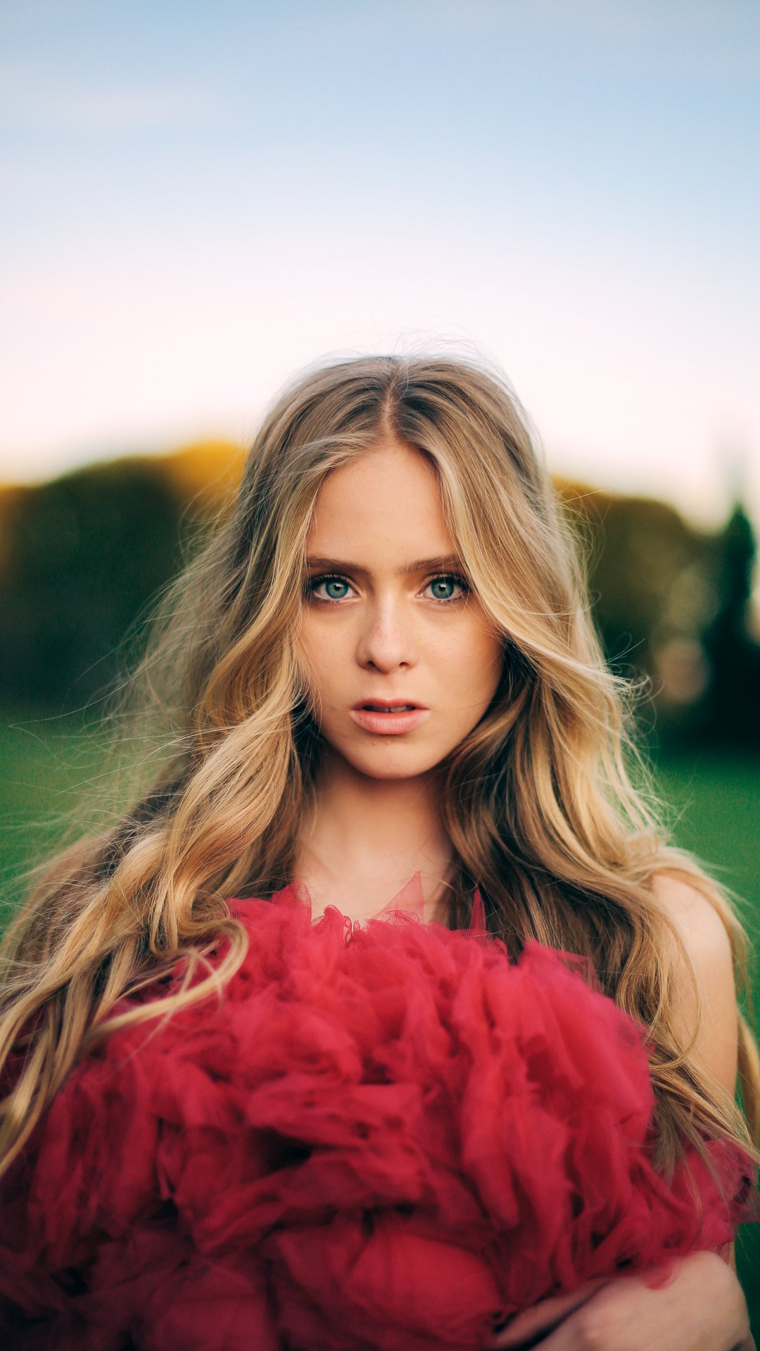 Young woman with long, wavy blonde hair and blue eyes holding a red, fluffy feathered object outdoors during sunset.