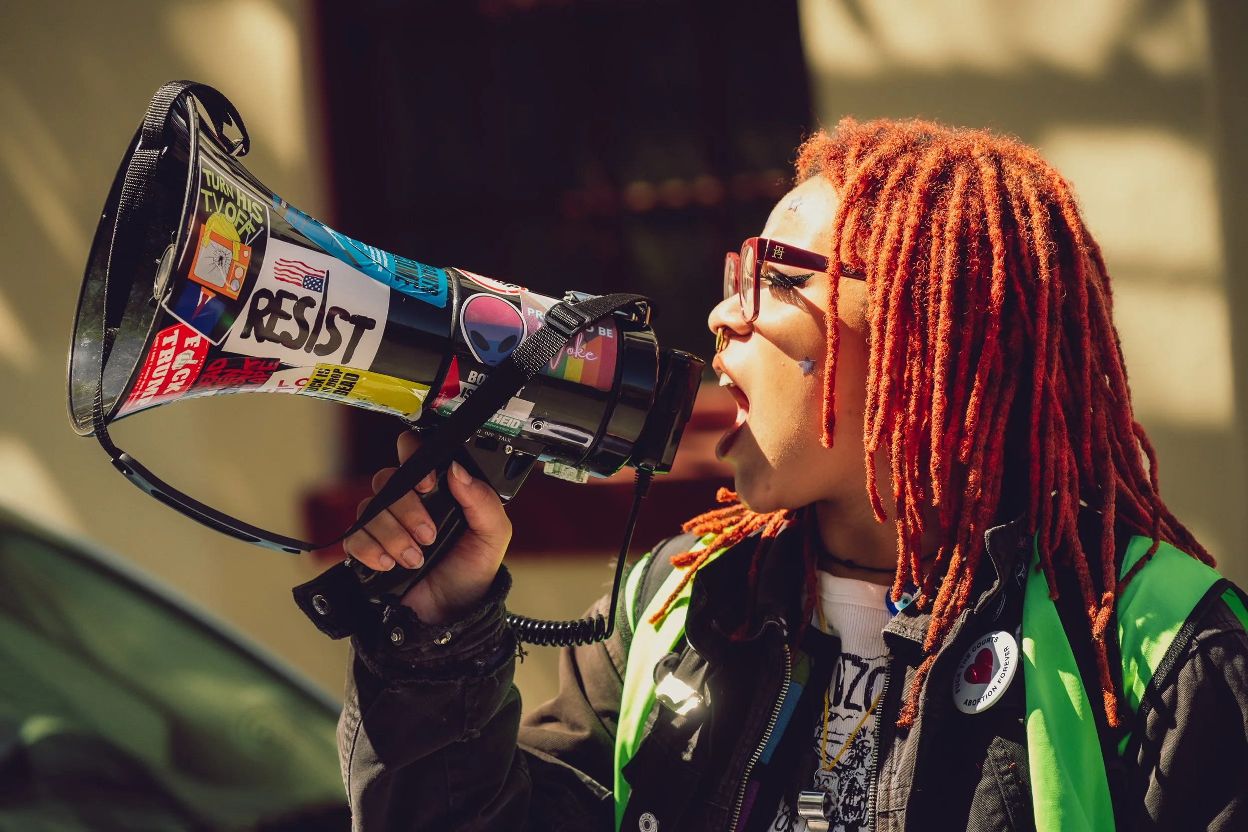 Person with red dreadlocks, wearing sunglasses and a green safety vest, shouting into a megaphone decorated with protest stickers.