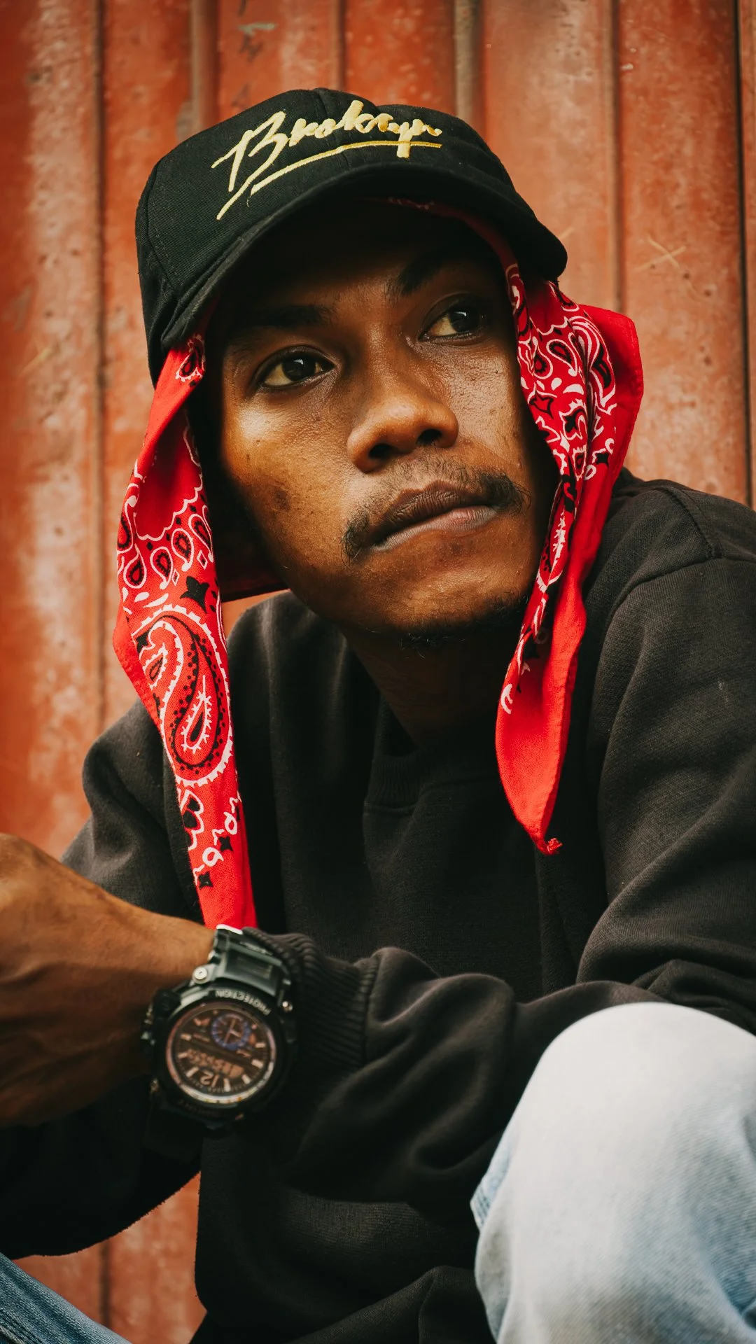 A young man wearing a black baseball cap with a gold logo, a red bandana tied around his head, a black sweatshirt, and a black watch, sitting against a rust-colored background.