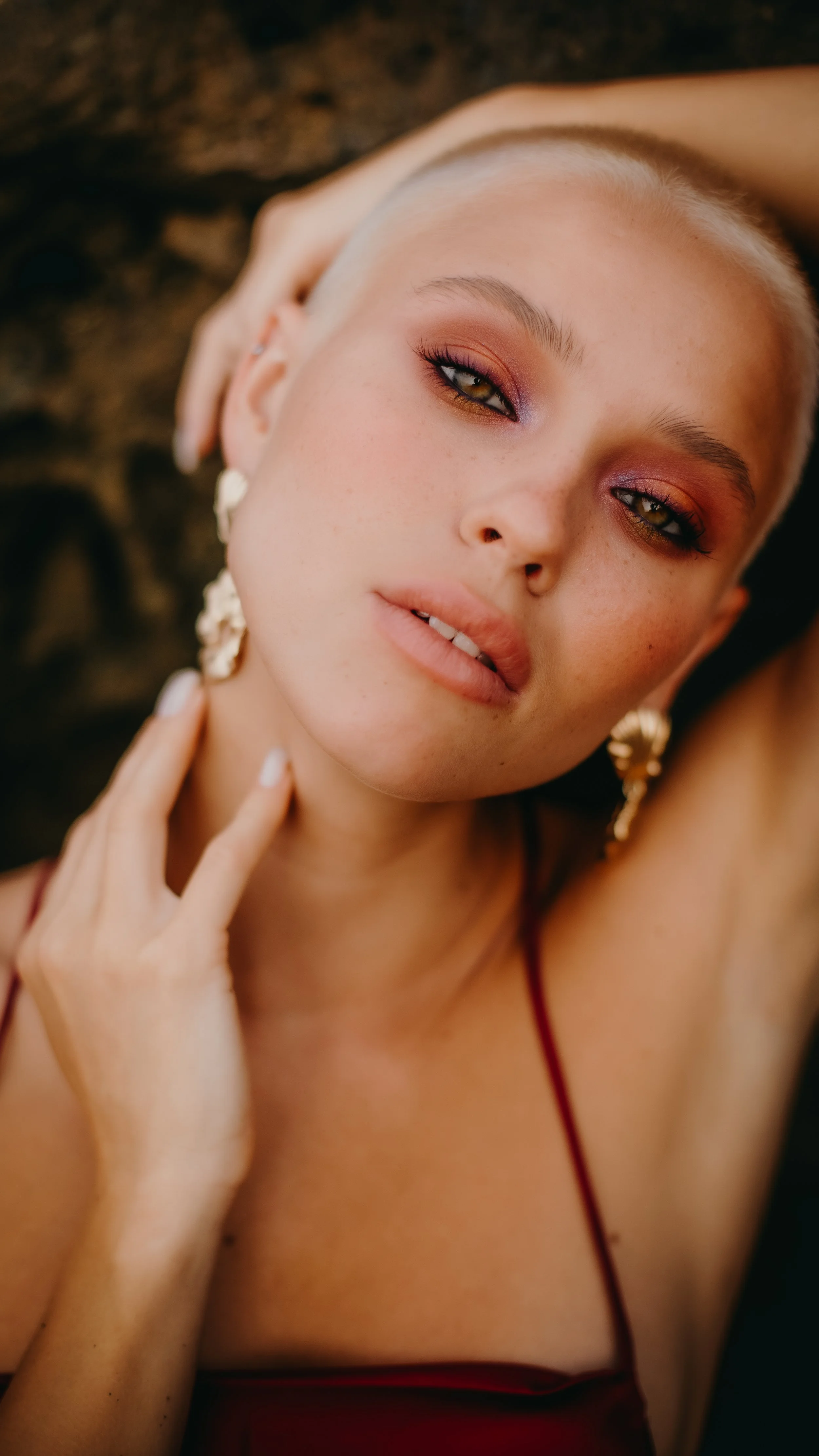 A close-up portrait of a woman with platinum blonde hair, wearing makeup with warm tones, earrings, and a red top, posing indoors.