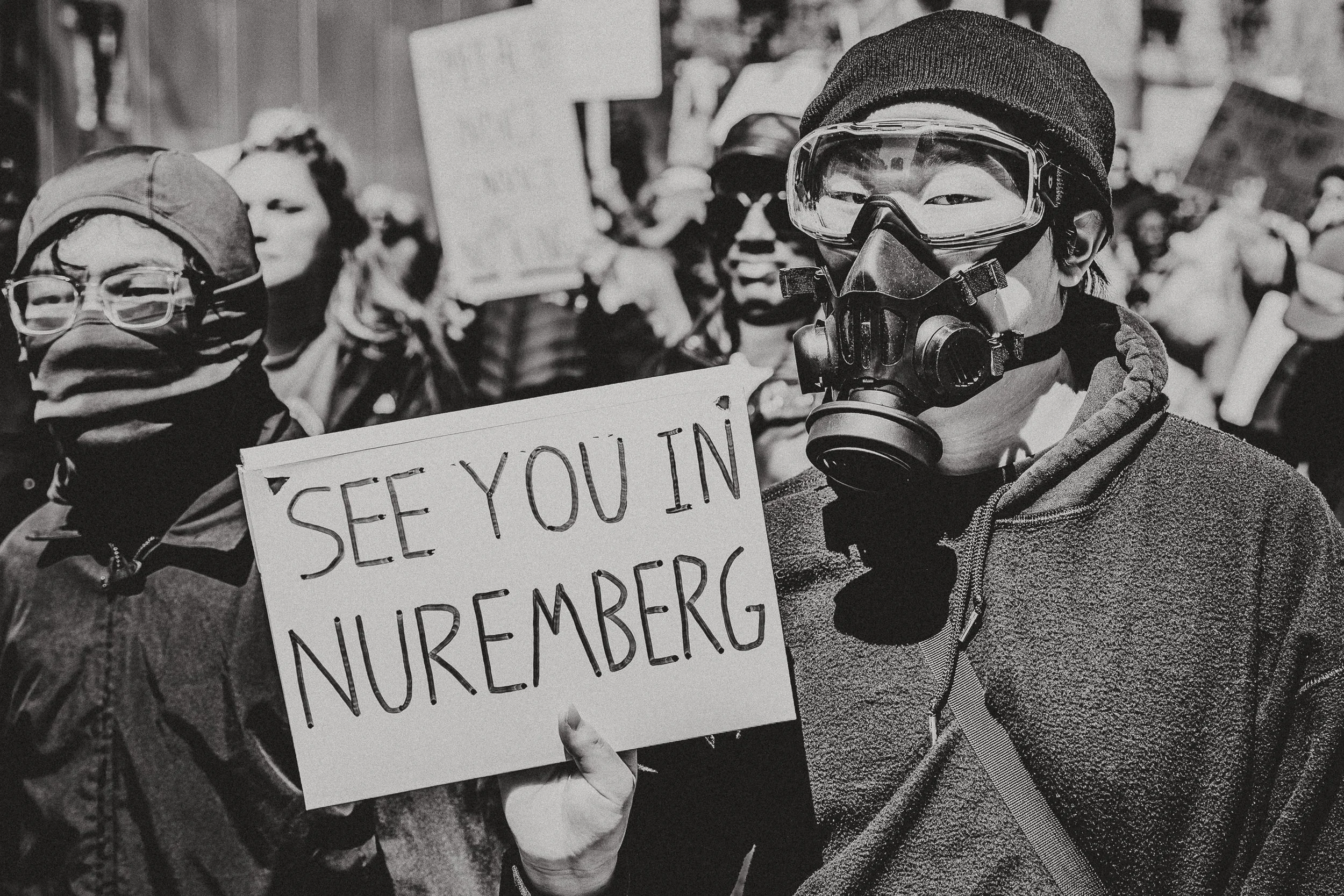 Protesters wearing masks and holding signs, with one person prominently holding a sign that reads 'SEE YOU IN NUREMBERG'.