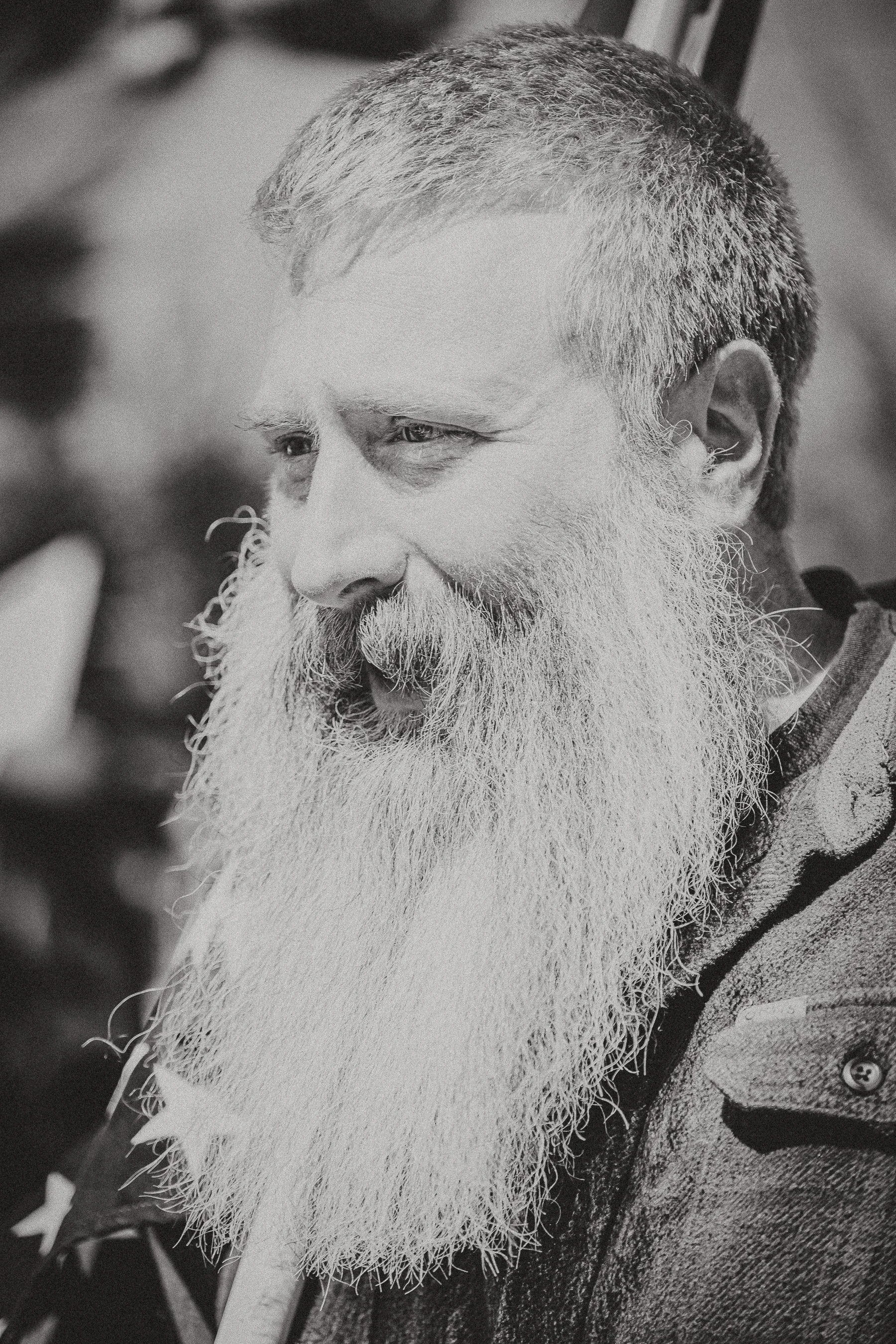 A black and white close-up of a smiling man with a long, thick beard and short hair, wearing a jacket.