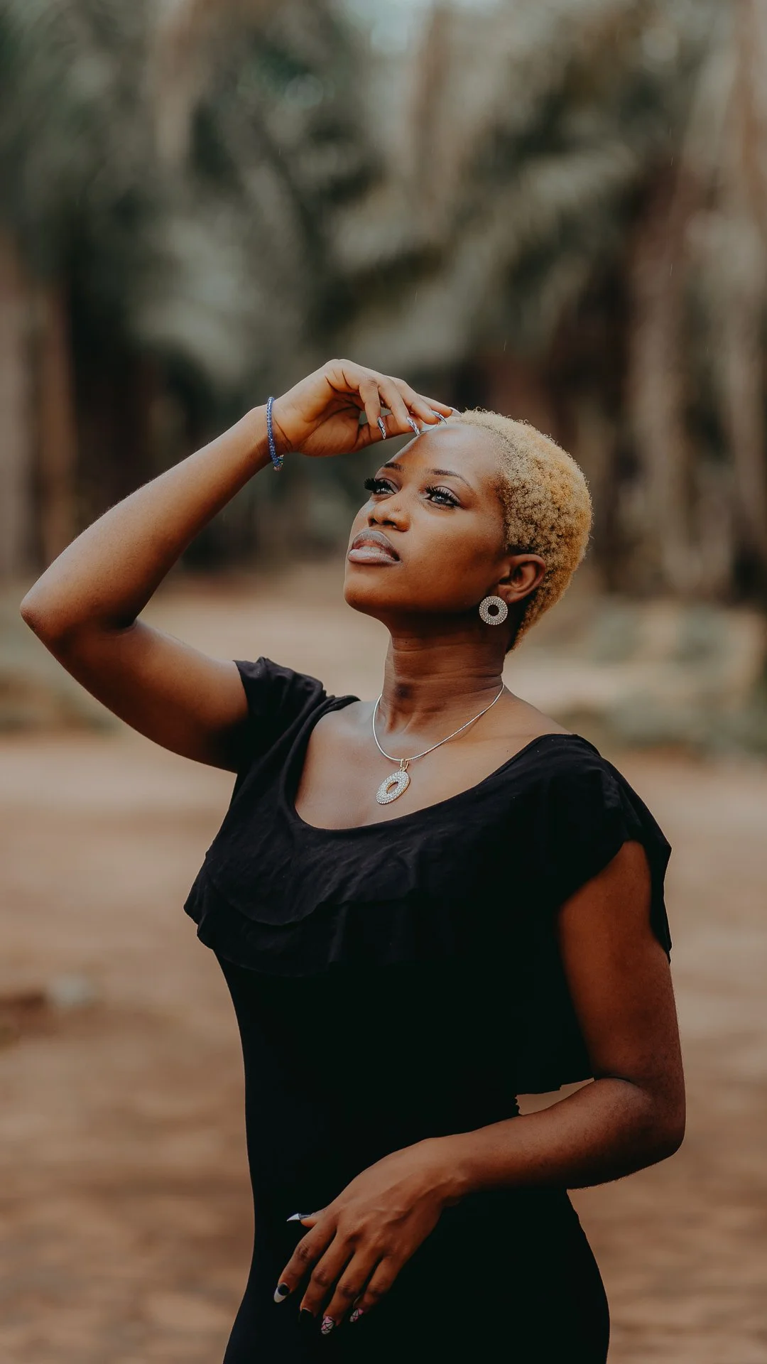 A woman with short, blonde hair wearing a black dress, jewelry, and standing outdoors with trees in the background.