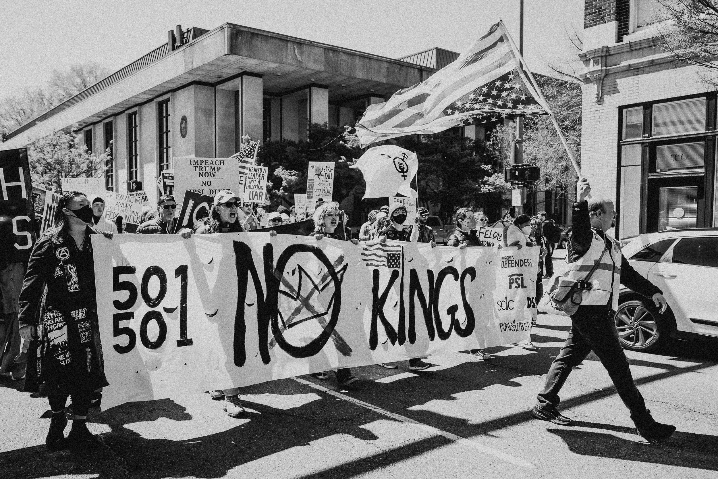 A black and white photo of a protest march with people holding signs and a large banner that reads '$501'. One person is carrying an American flag. The protesters appear to be advocating against corporate greed, with signs mentioning impeachment, lyi