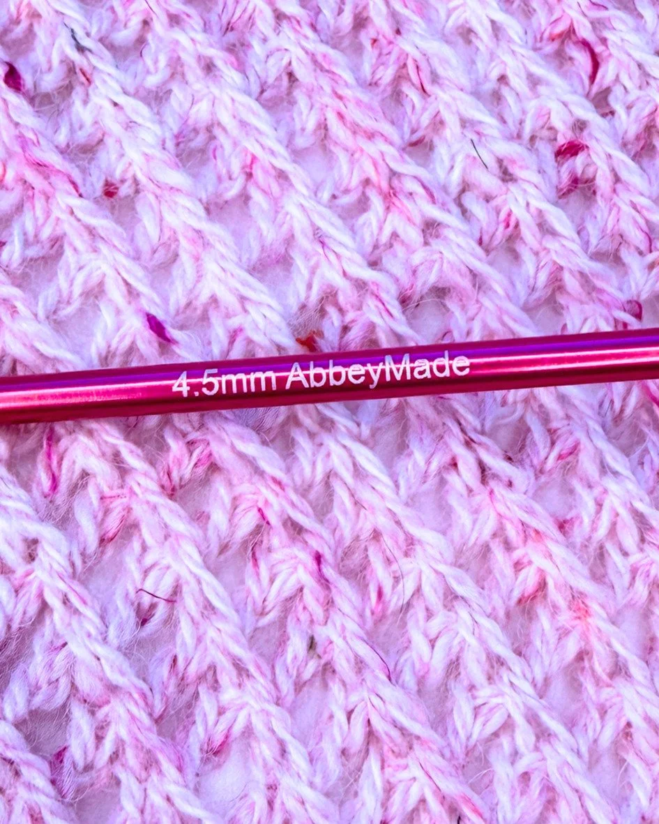 Close-up of a pink Tunisian crochet hook with '4.5 mm AbbeyMade written on it, resting on a pink and white textured Tunisian crochet fabric.