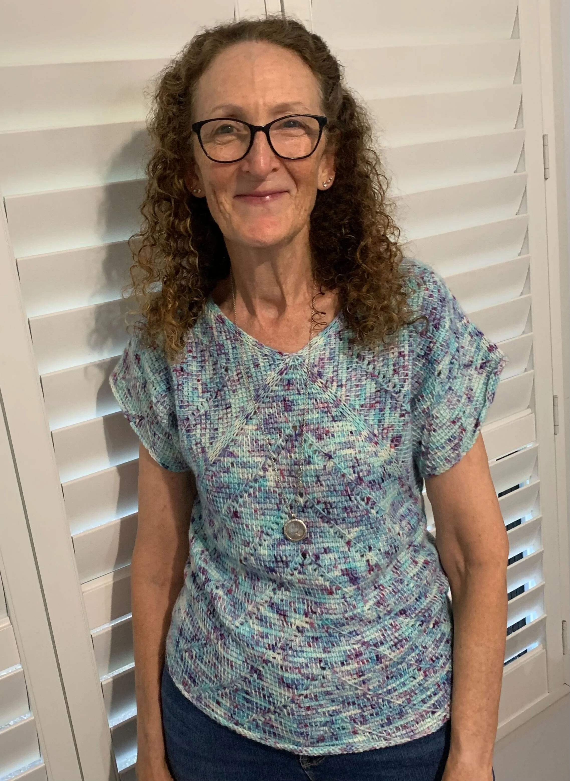 A smiling elderly woman with curly hair and glasses, wearing a multicolored patterned top, standing in front of white plantation shutters.